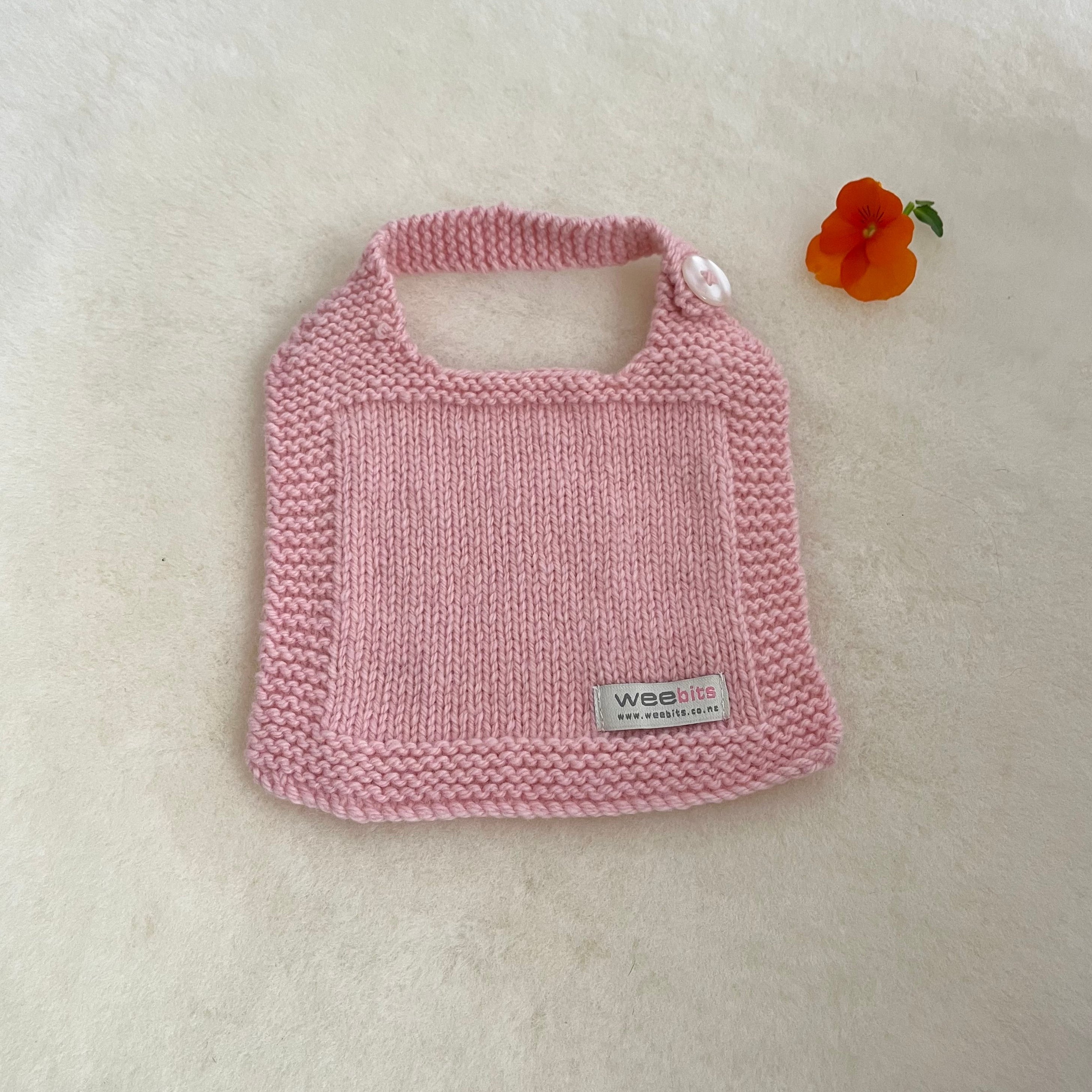A pink hand-knit baby bib with a small white pearl button and a small woven label reading 'weebits', displayed on a soft white textured background next to a small orange flower.