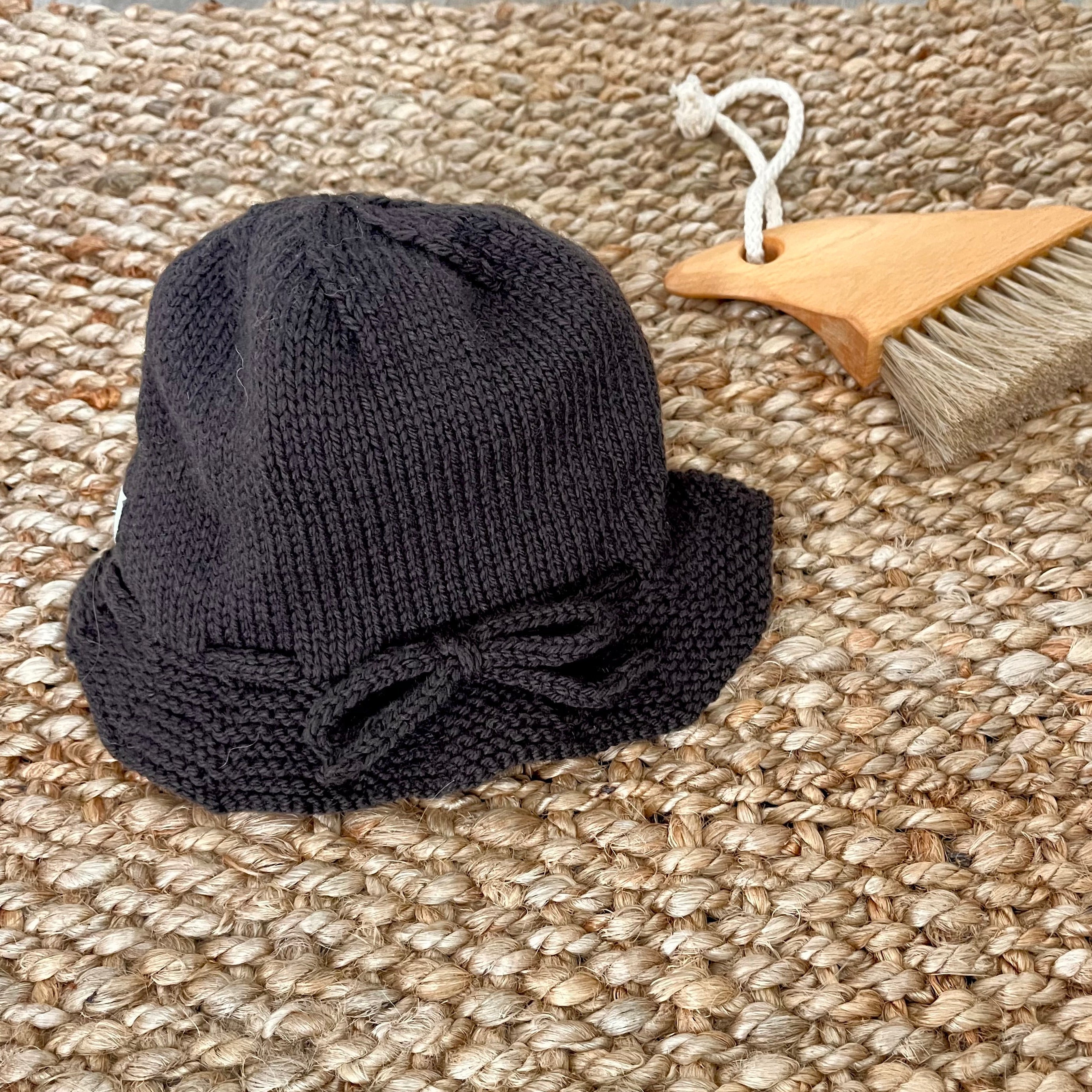 A brown knit baby hat with a small bow on the brim, displayed on a woven jute rug next to a wooden hairbrush.