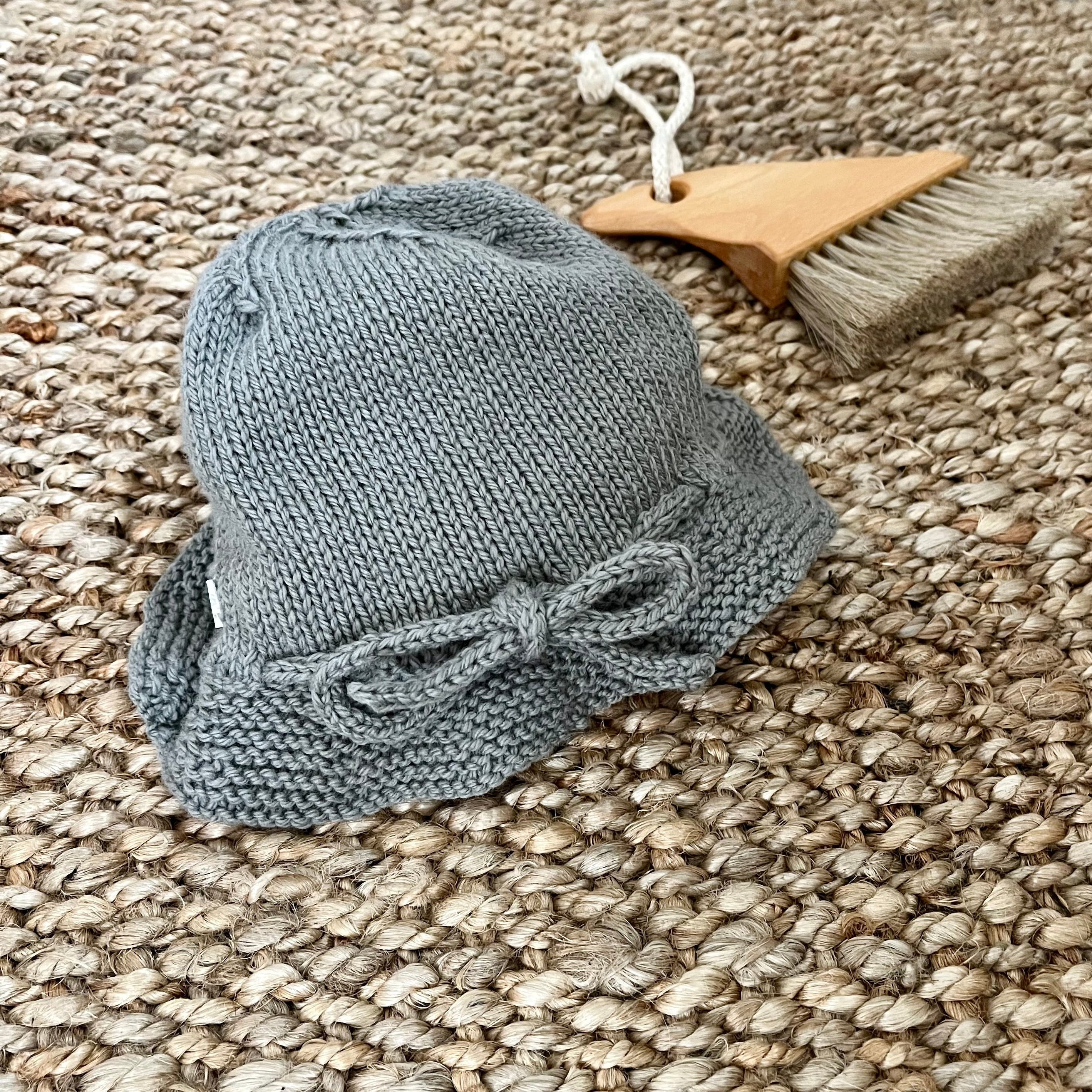 A small gray knitted bucket hat with a bow detail, displayed on a woven jute rug next to a wooden baby brush.
