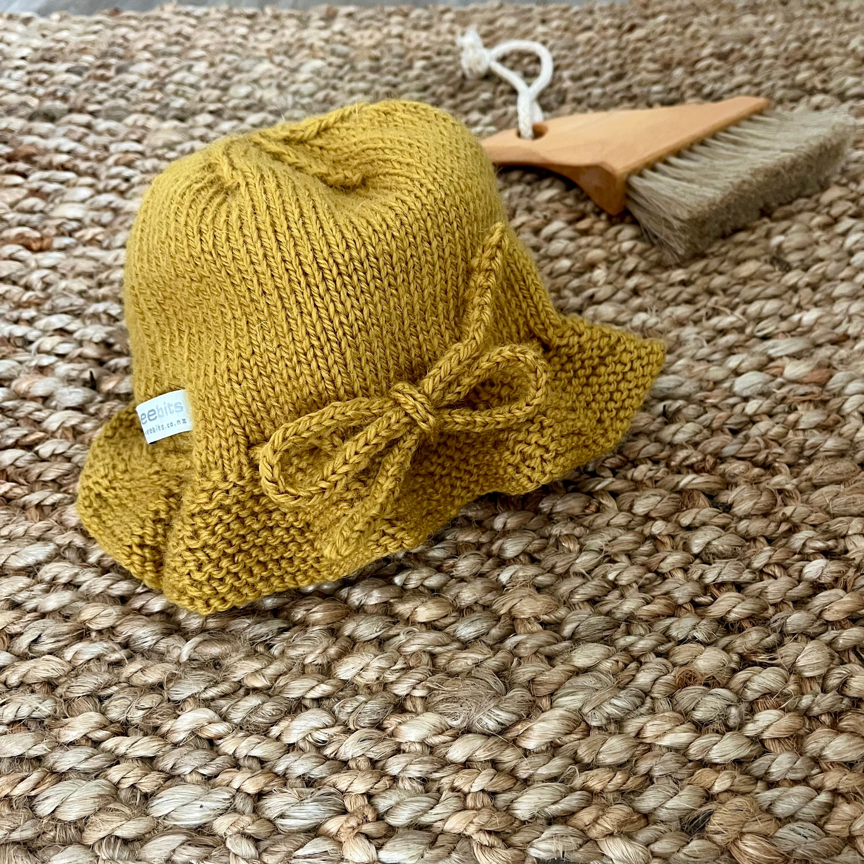 A small mustard knitted bucket hat with a bow detail, displayed on a woven jute rug next to a wooden beach brush.