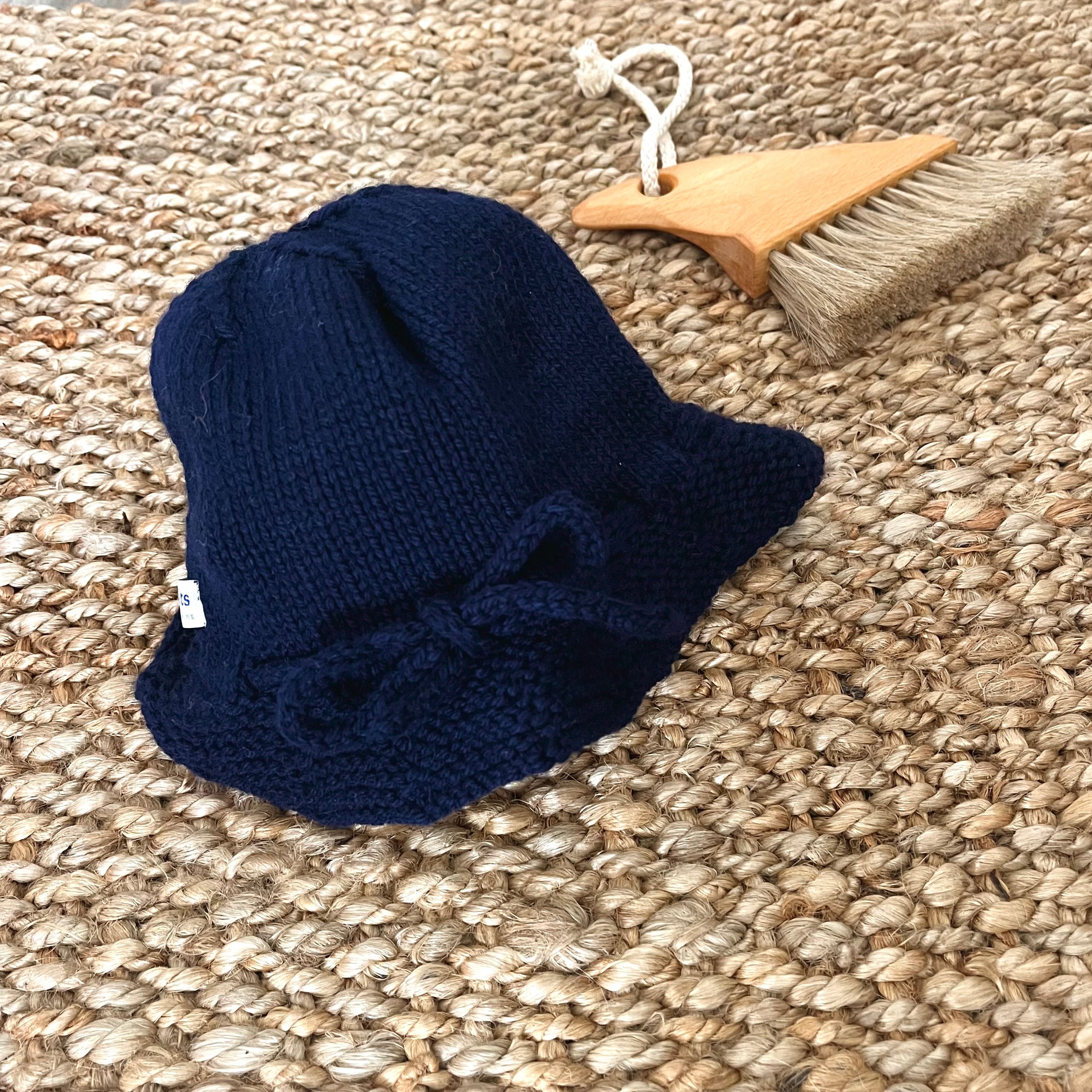 A small navy knitted bucket hat with a bow detail, displayed on a woven jute rug next to a wooden beach brush.