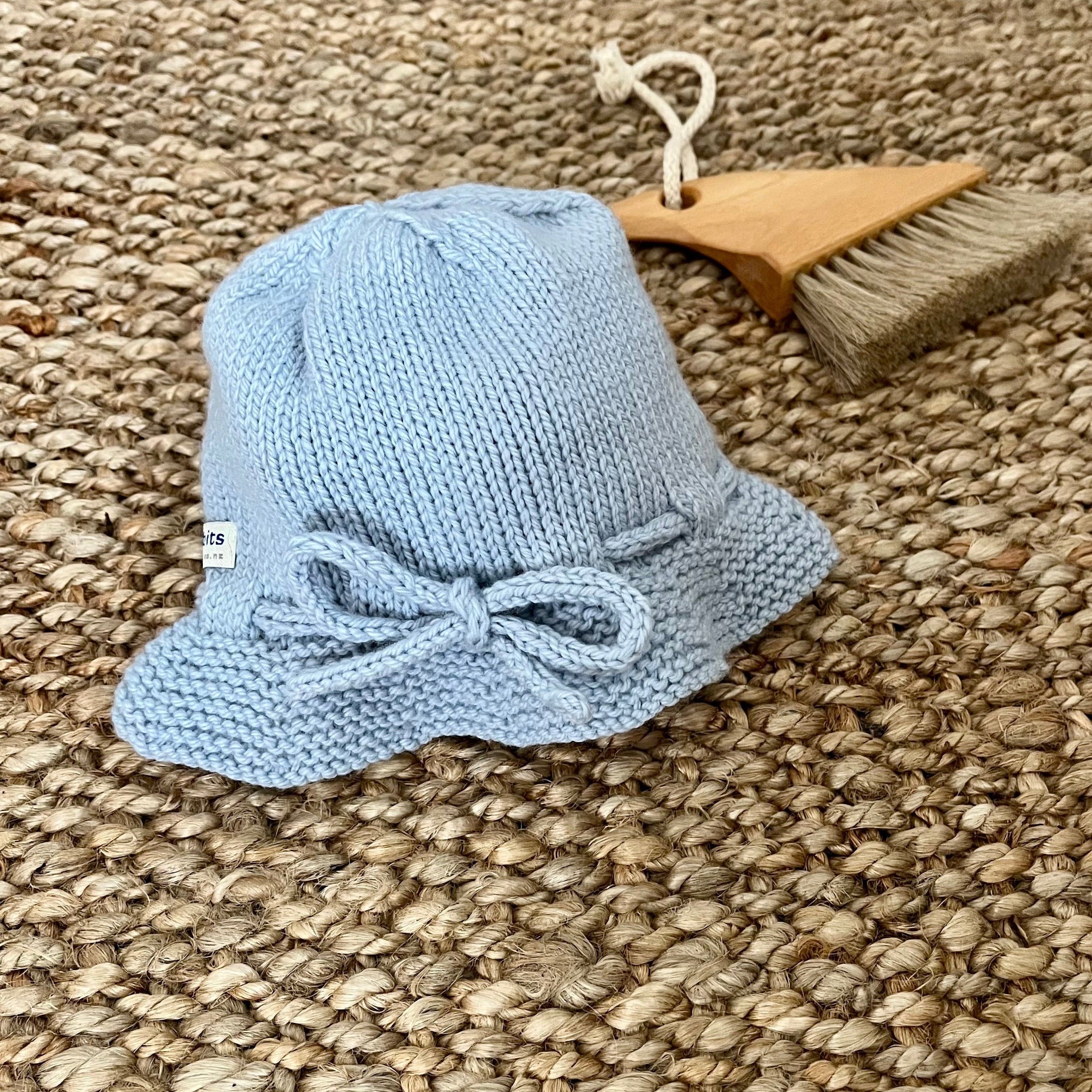 A small blue knitted bucket hat with a bow detail, displayed on a woven jute rug next to a wooden beach brush.