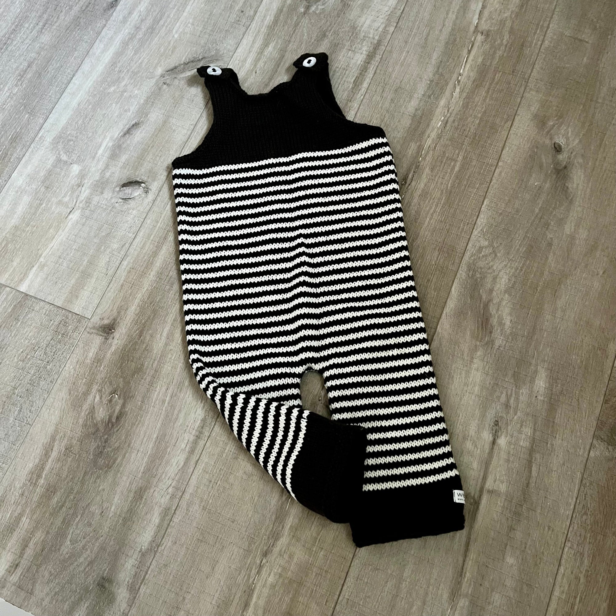 A chocolate natural stripe colored knit baby dungaree with shoulder straps and a leg opening, laid flat on a light brown wood floor.