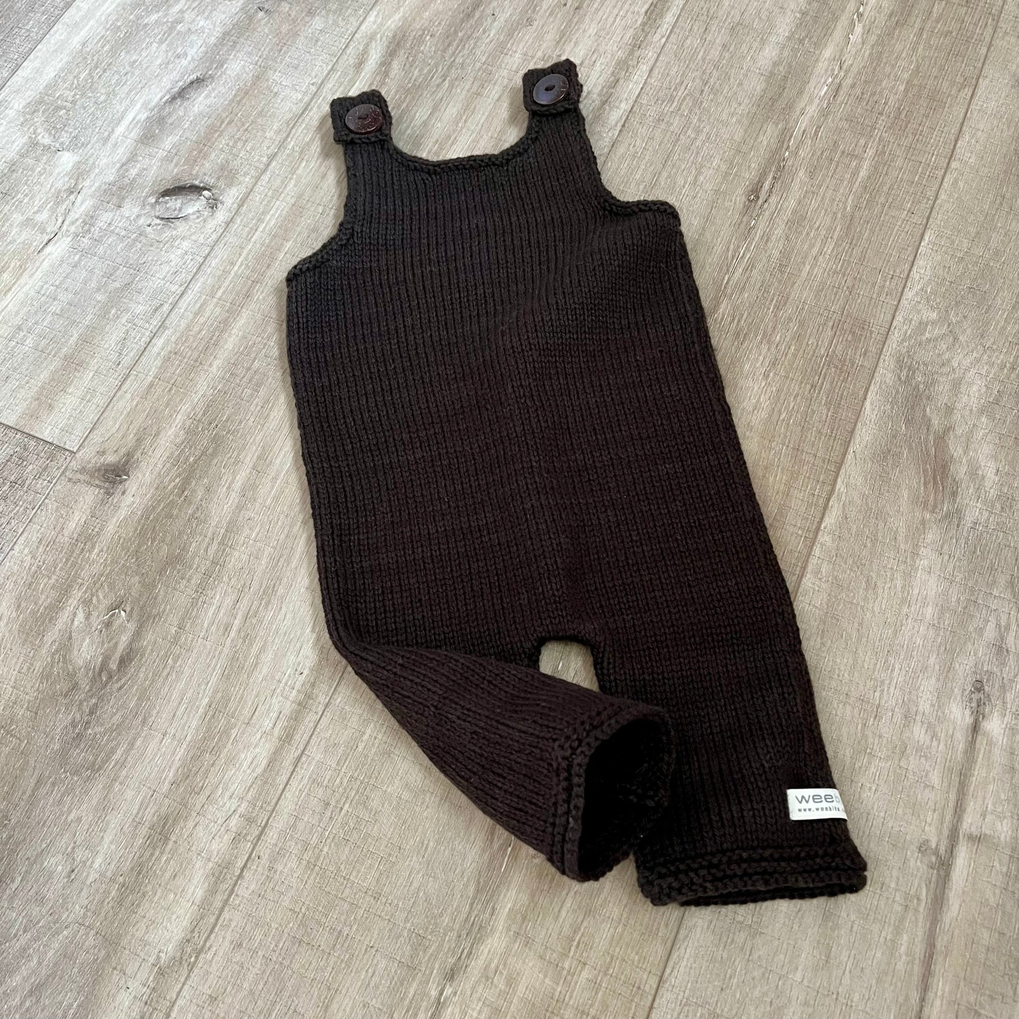 A dark brown ribbed knit baby romper with button shoulder straps, laid flat on a light wood plank floor. A small white brand tag is visible on the lower leg.