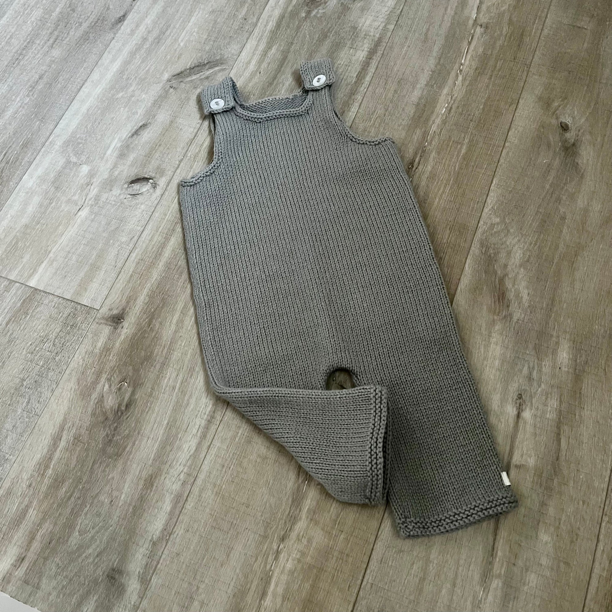 A mushroom colored knit baby dungaree with shoulder straps and a leg opening, laid flat on a light brown wood floor.