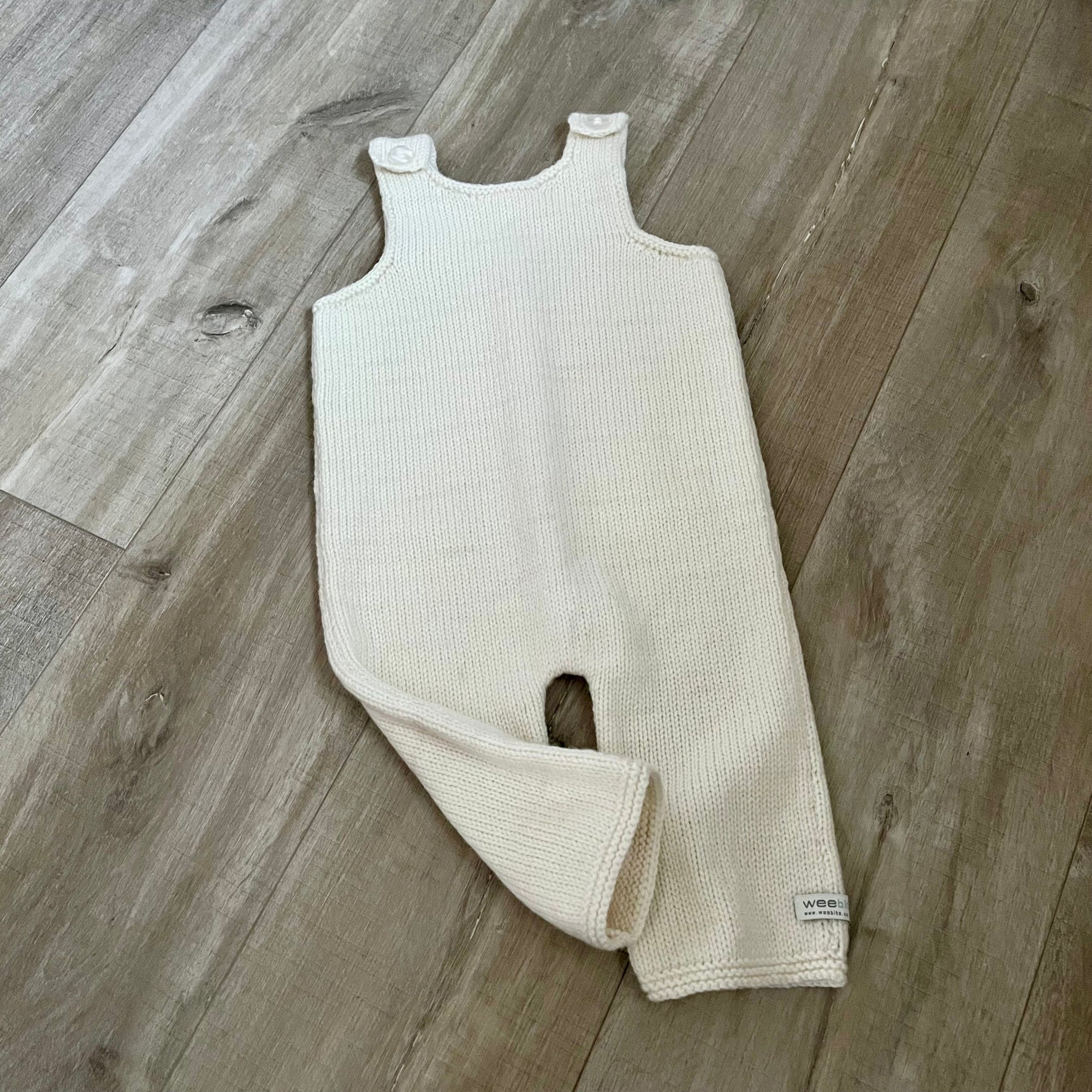 A natural colored knit baby dungaree with shoulder straps and a leg opening, laid flat on a light brown wood floor.