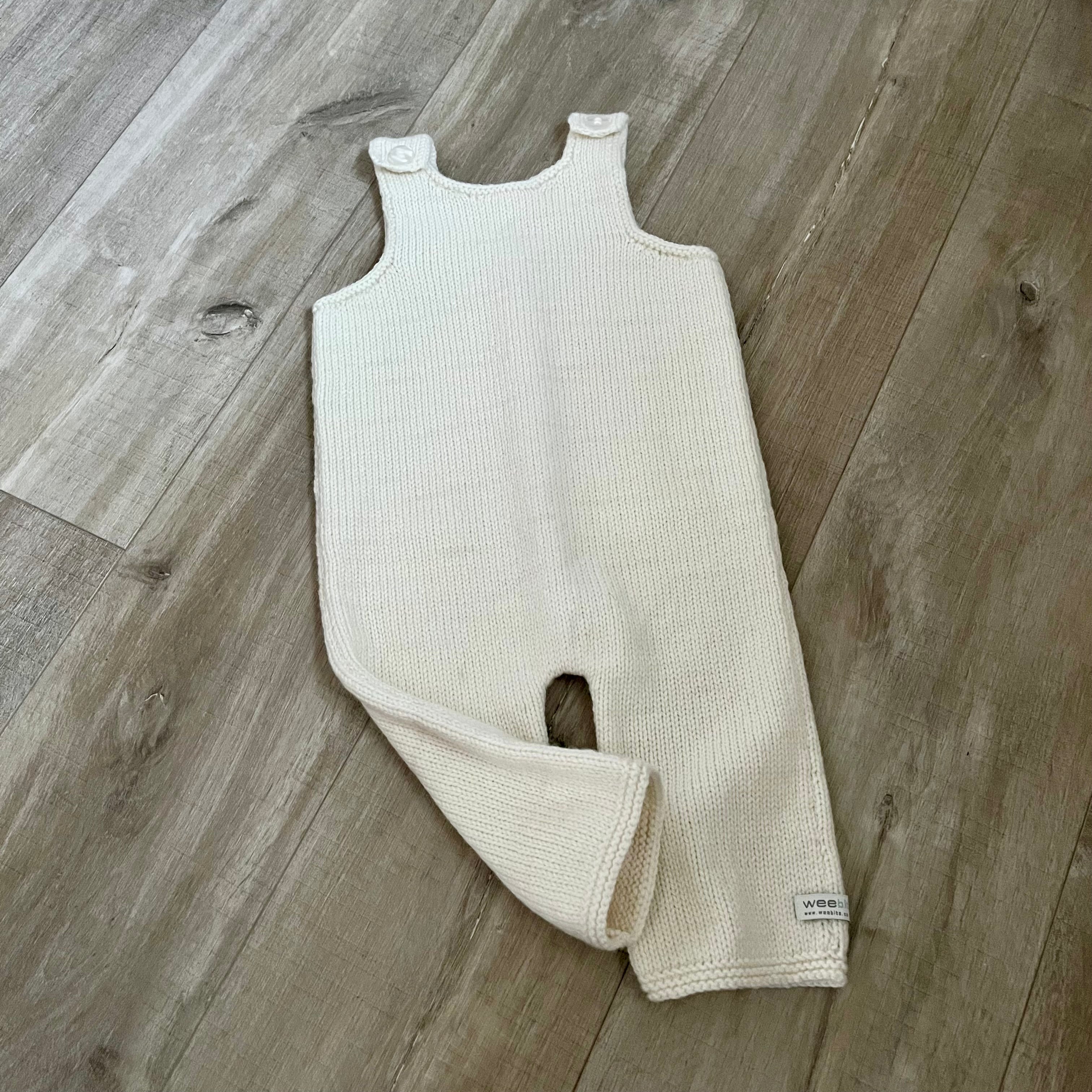 A natural colored knit baby dungaree with shoulder straps and a leg opening, laid flat on a light brown wood floor.