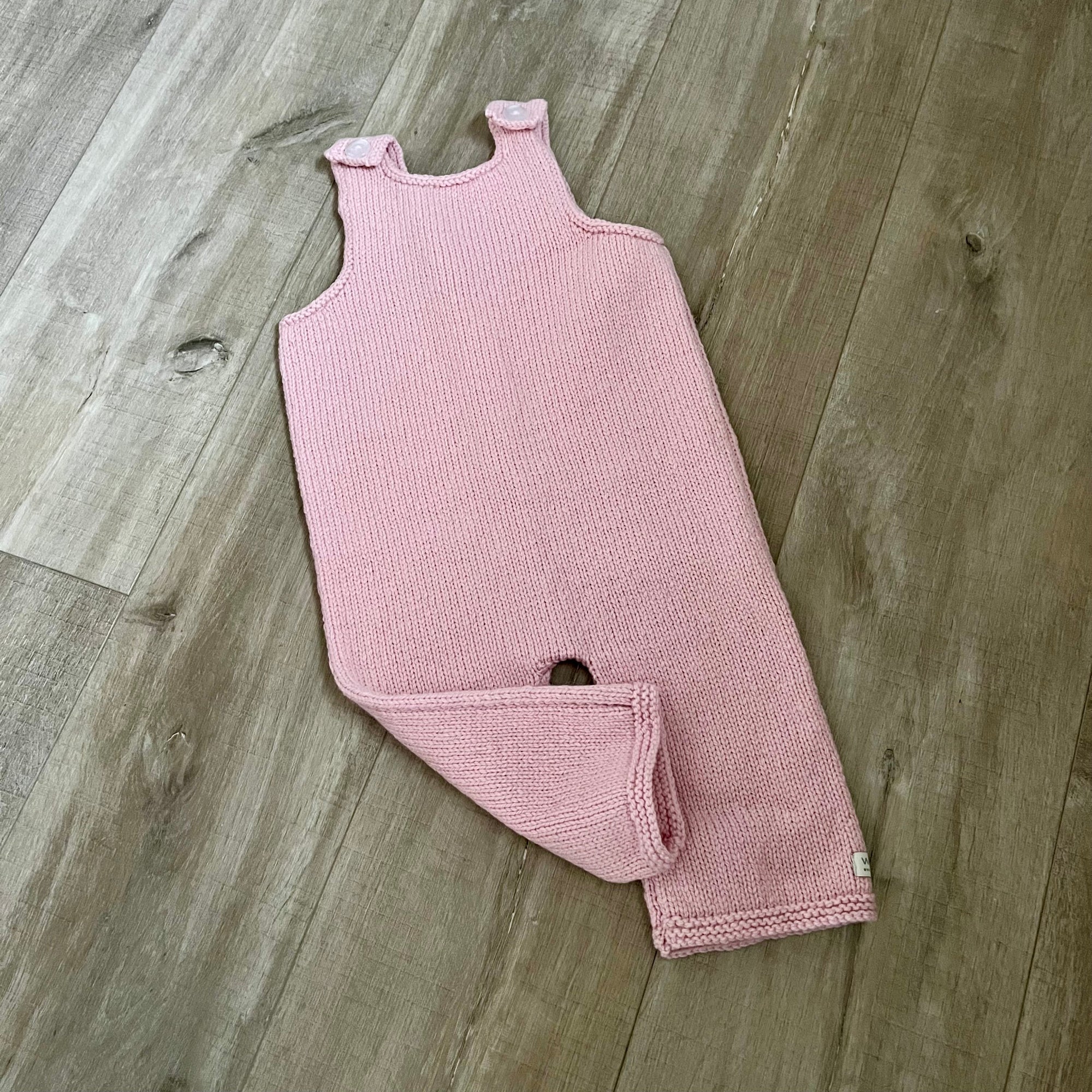 A pink colored knit baby dungaree with shoulder straps and a leg opening, laid flat on a light brown wood floor.