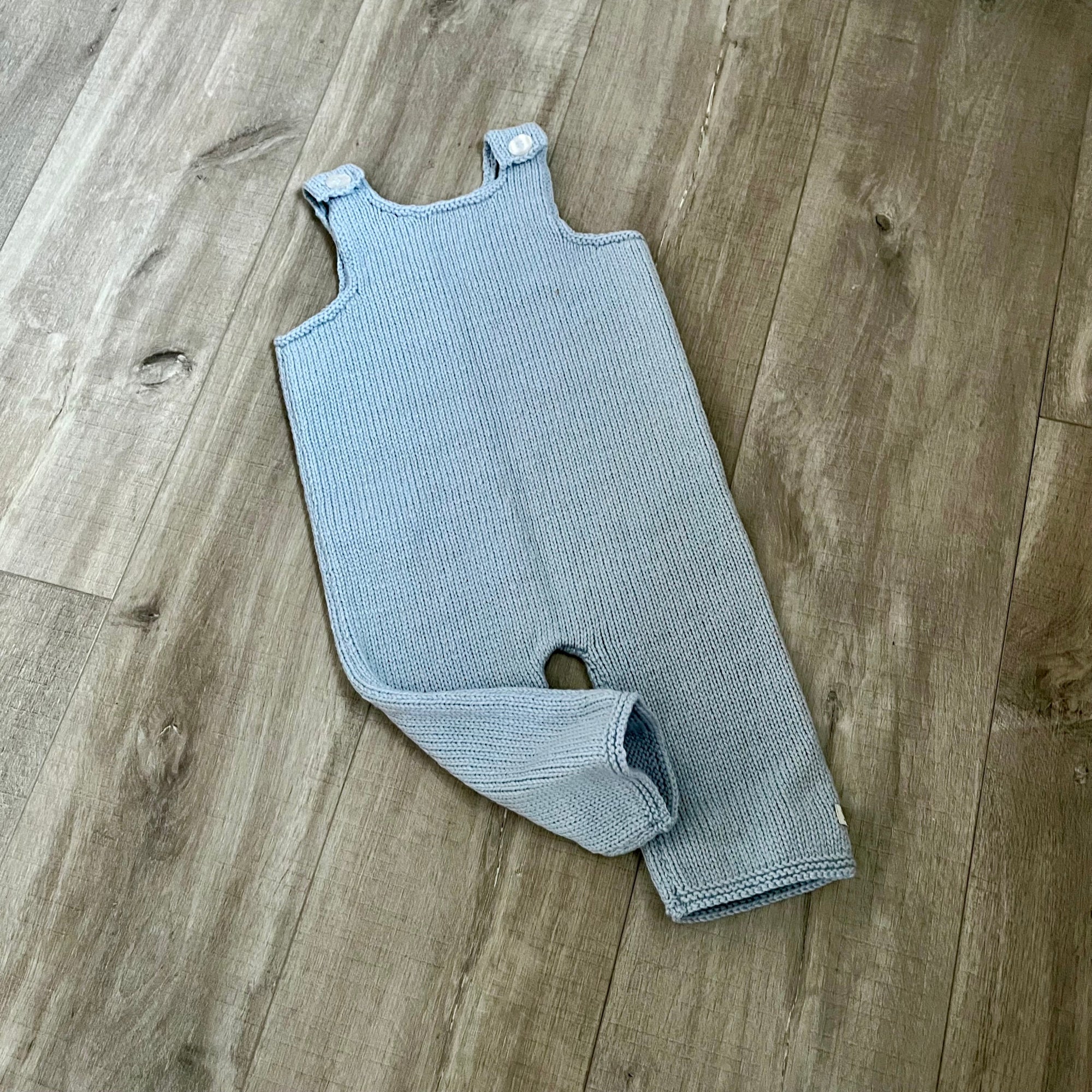 A blue colored knit baby dungaree with shoulder straps and a leg opening, laid flat on a light brown wood floor.