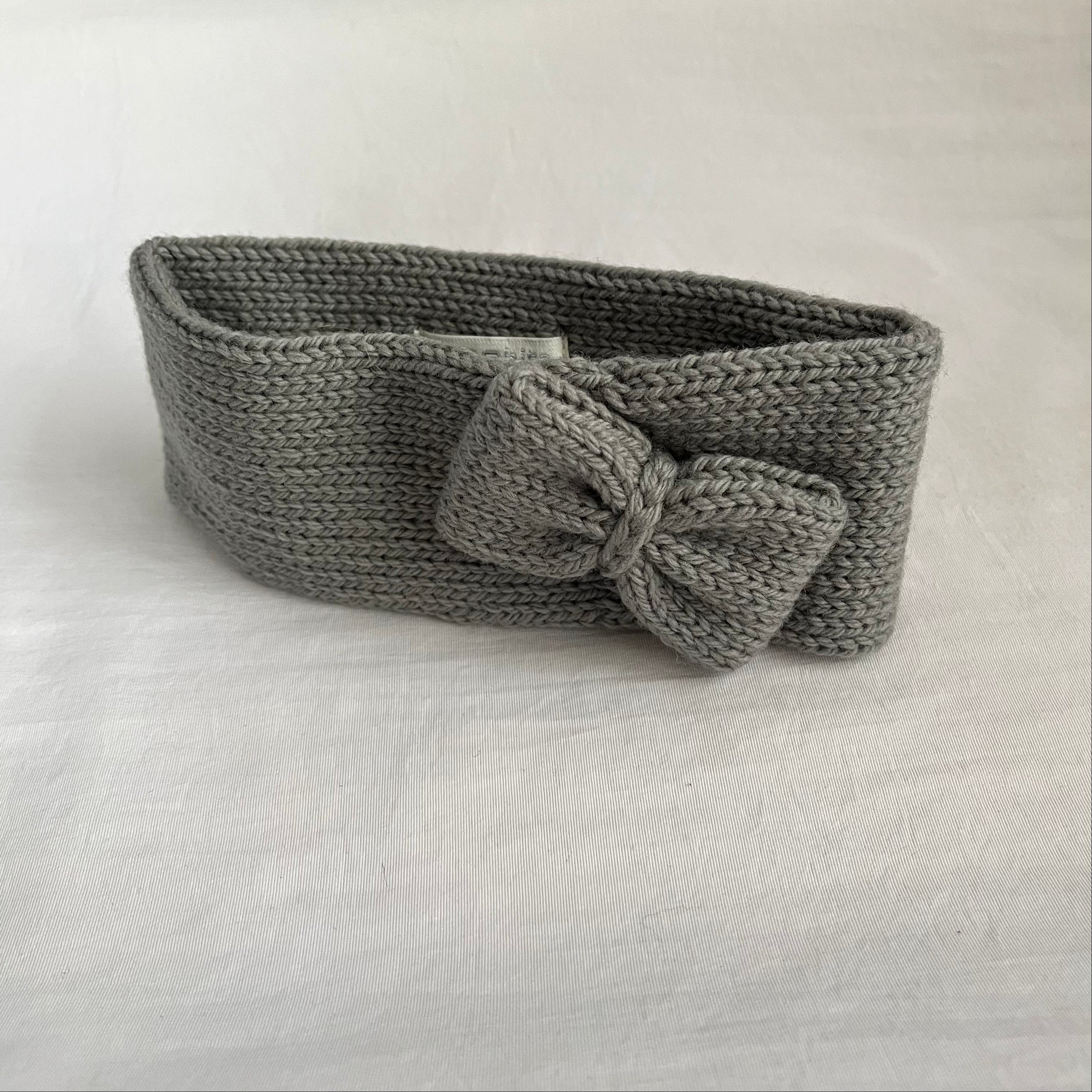 A gray knitted headband with a large bow detail, displayed on a light gray fabric background.