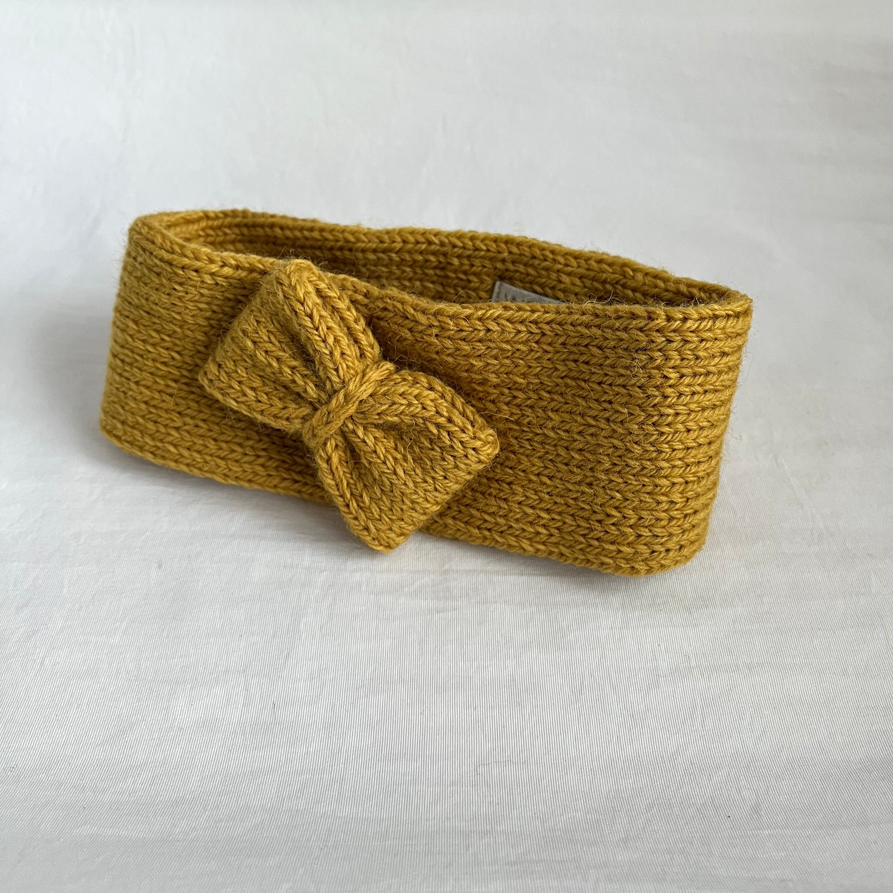 A wide, chunky knit headband in a warm mustard yellow color, featuring a large matching bow on the front, displayed on a textured white fabric background.