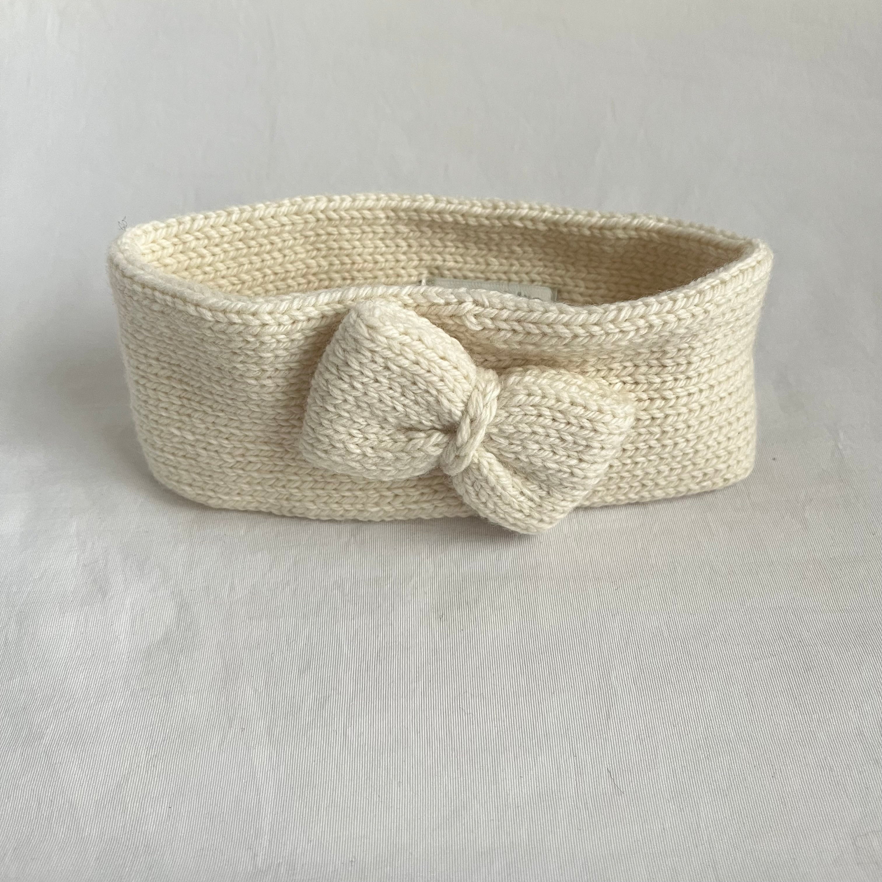 A cream-colored, hand-knit headband with a large matching bow on the front, displayed on a textured light gray fabric background.