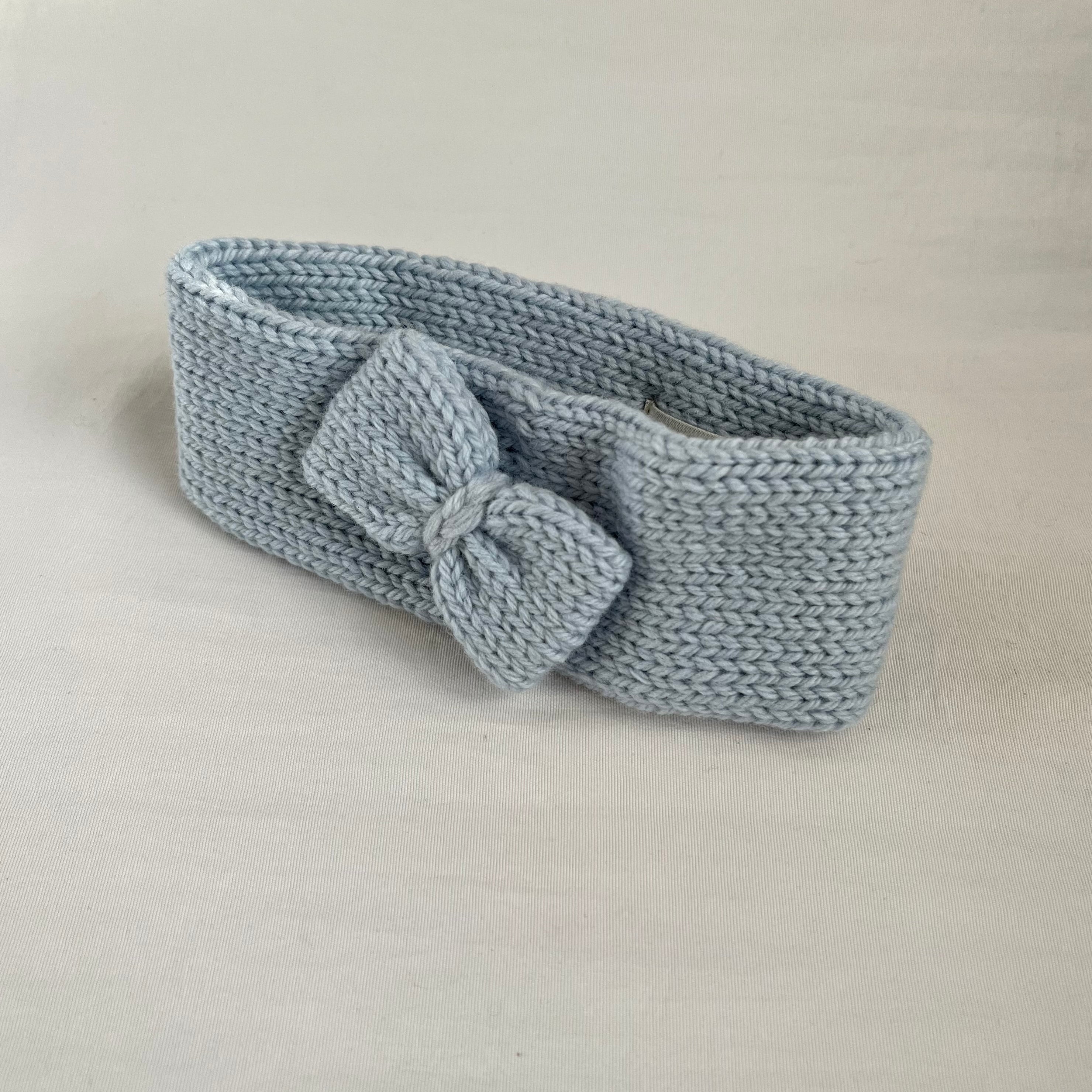 A light blue-gray knitted headband with a matching bow, displayed on a neutral light beige background.