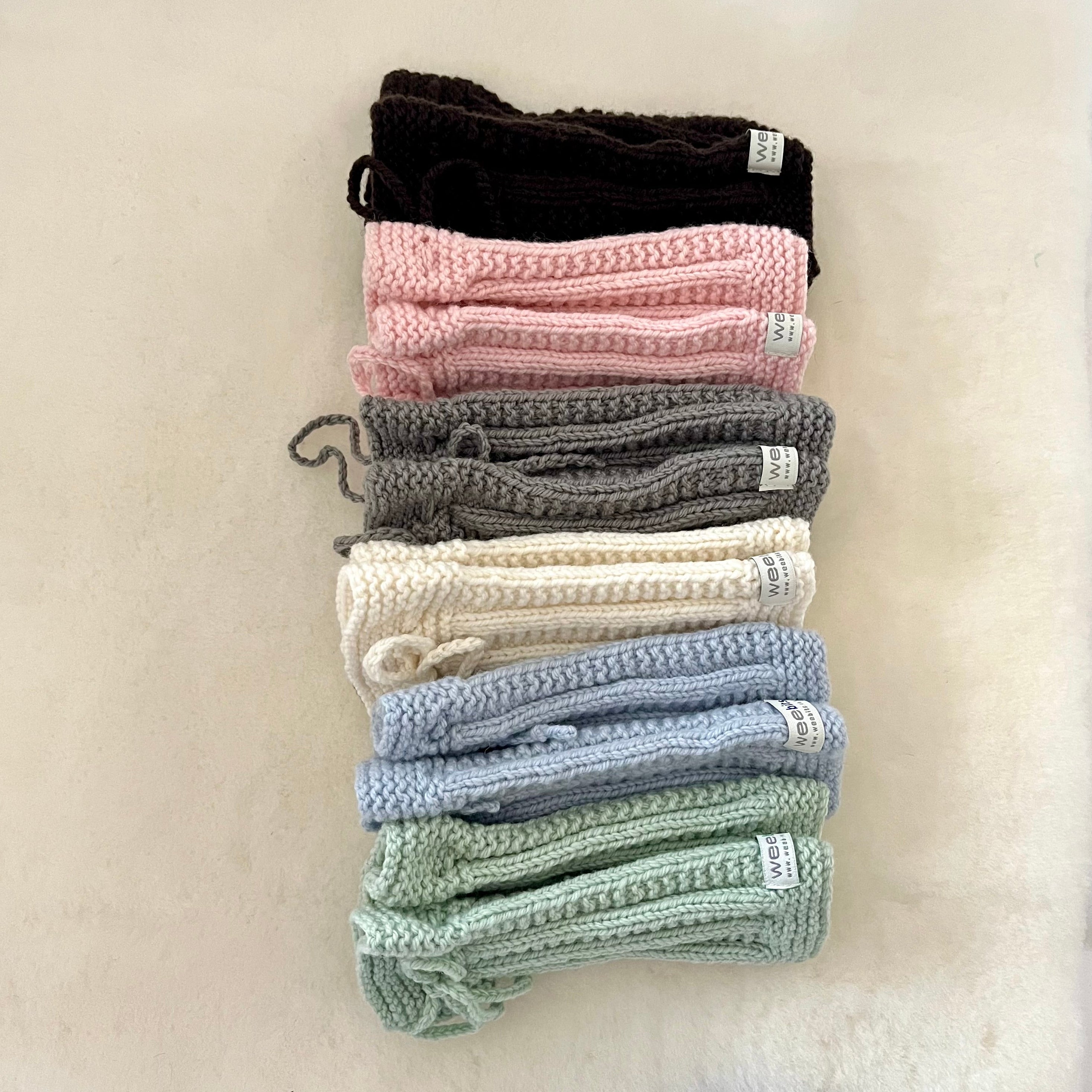 A stack of knitted legwarmers  in various colors, including black, pink, gray, cream, light blue, and mint green, arranged vertically on a soft off-white background.