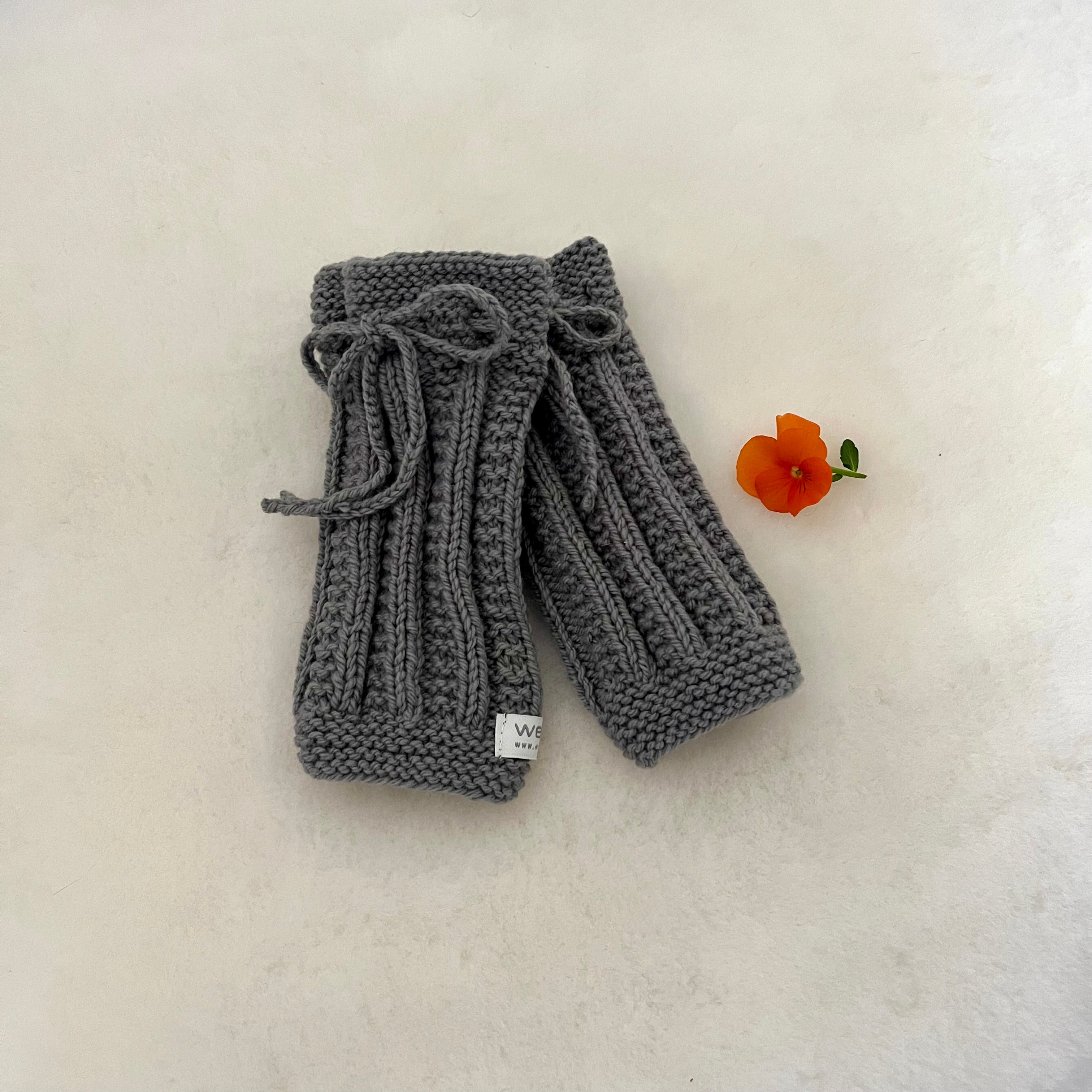 A pair of mushroom hand-knitted legwarmers with a matching knitted bow on the top, displayed on a textured off-white surface next to a small orange flower.