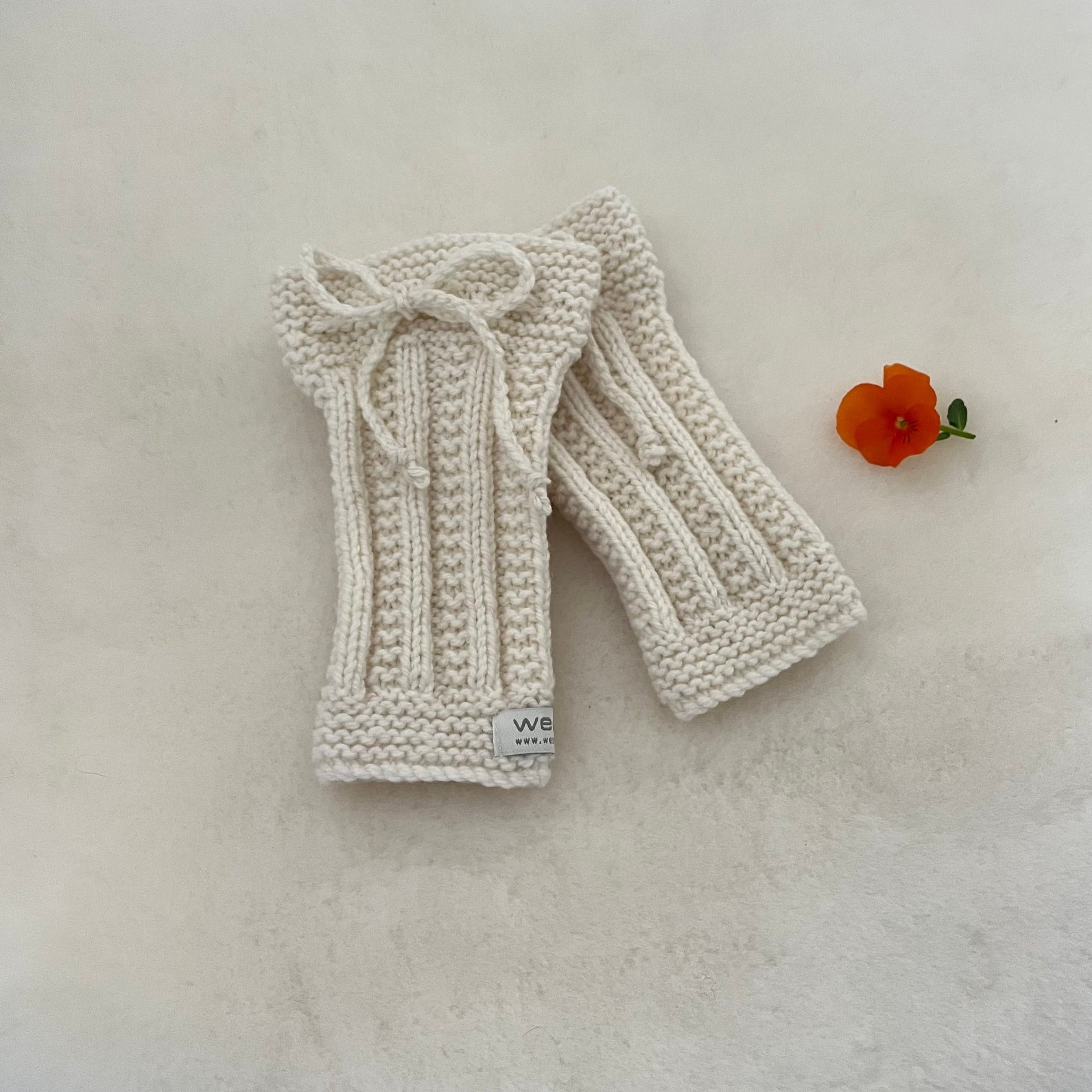 A pair of natural hand-knitted legwarmers with a matching knitted bow on the top, displayed on a textured off-white surface next to a small orange flower.