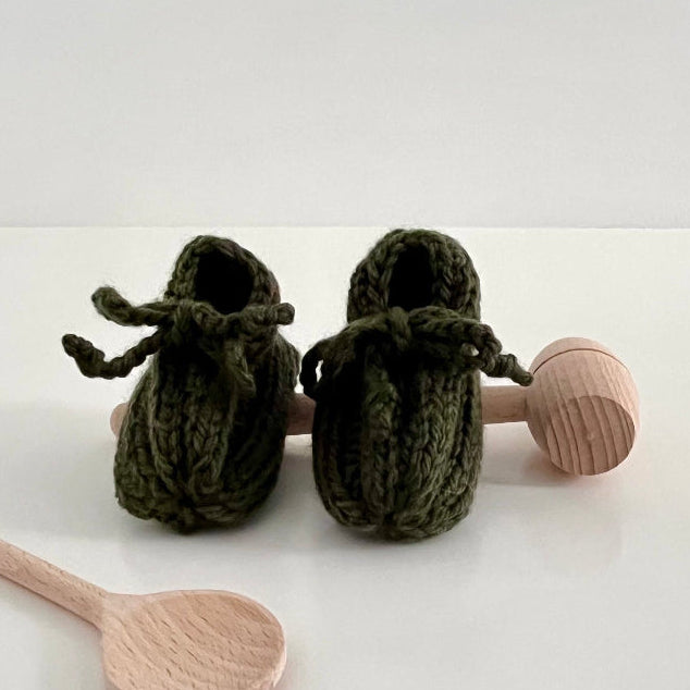 Forest green knitted baby booties with wooden rattle and spoon on a white background