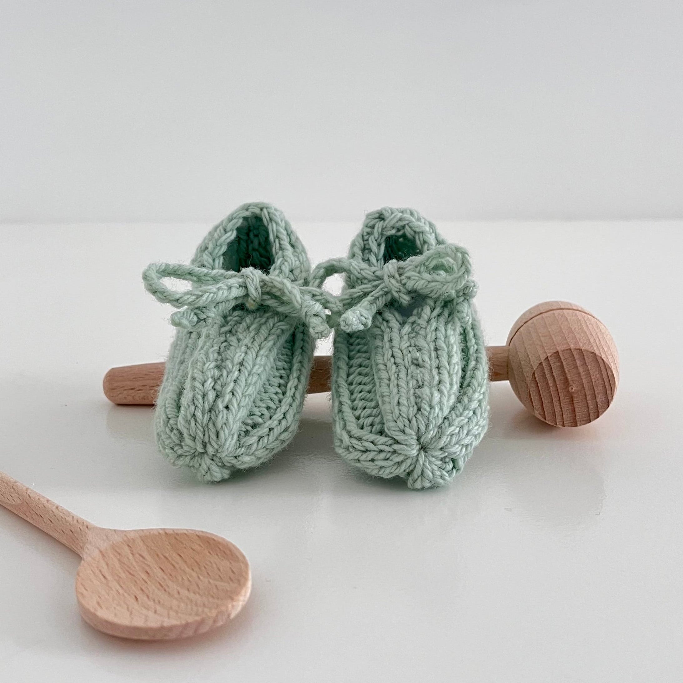 Pair of mint knitted baby shoes with wooden rattle and spoon on a white background