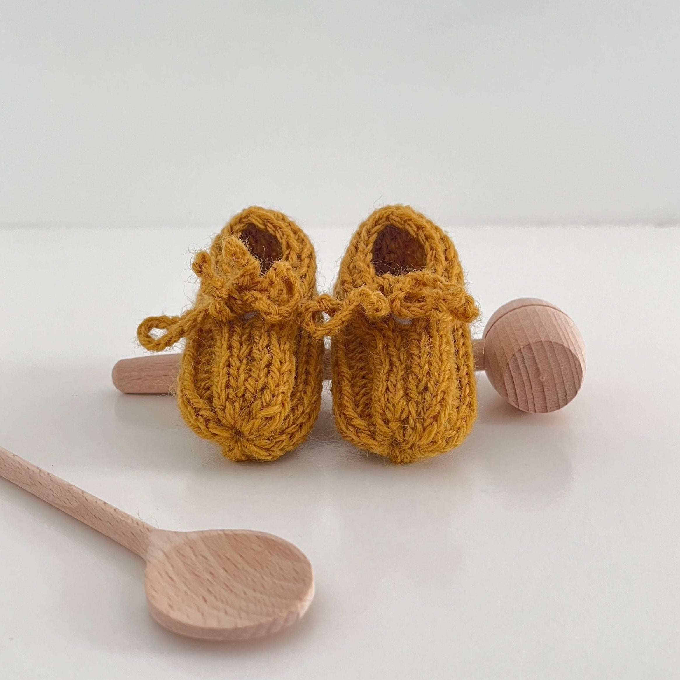 Pair of mustard knitted baby booties on a white background with wooden spoons for scale.