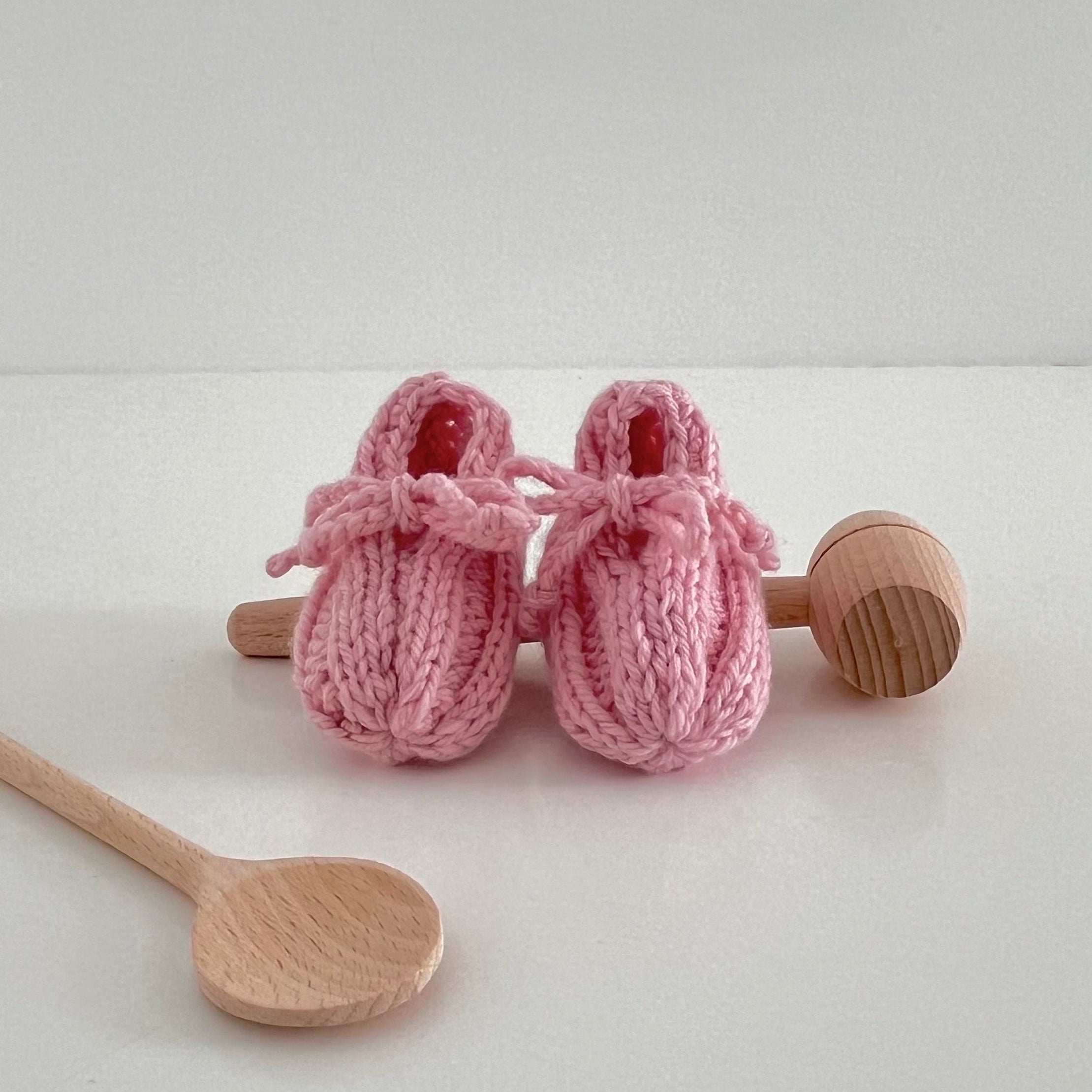 Pair of pink knitted baby booties with wooden spoons on a white background