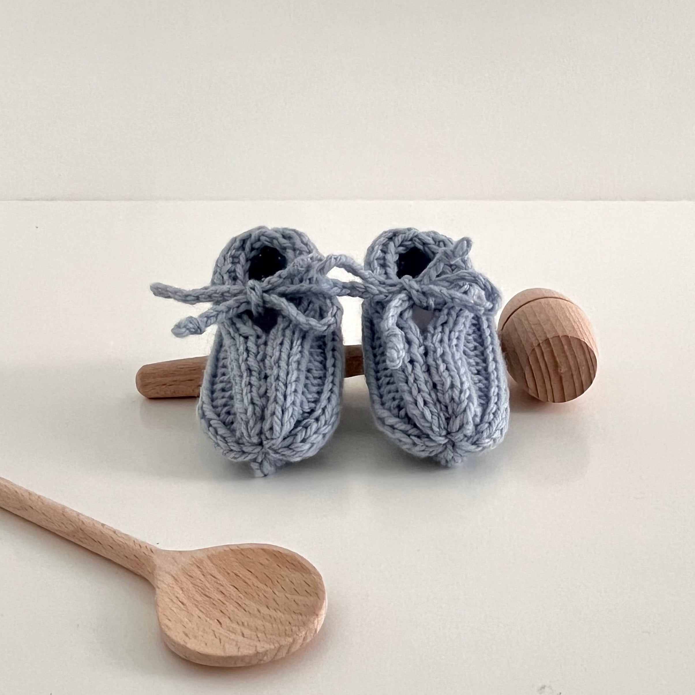 Pair of blue knitted baby booties with wooden spoons on a white background