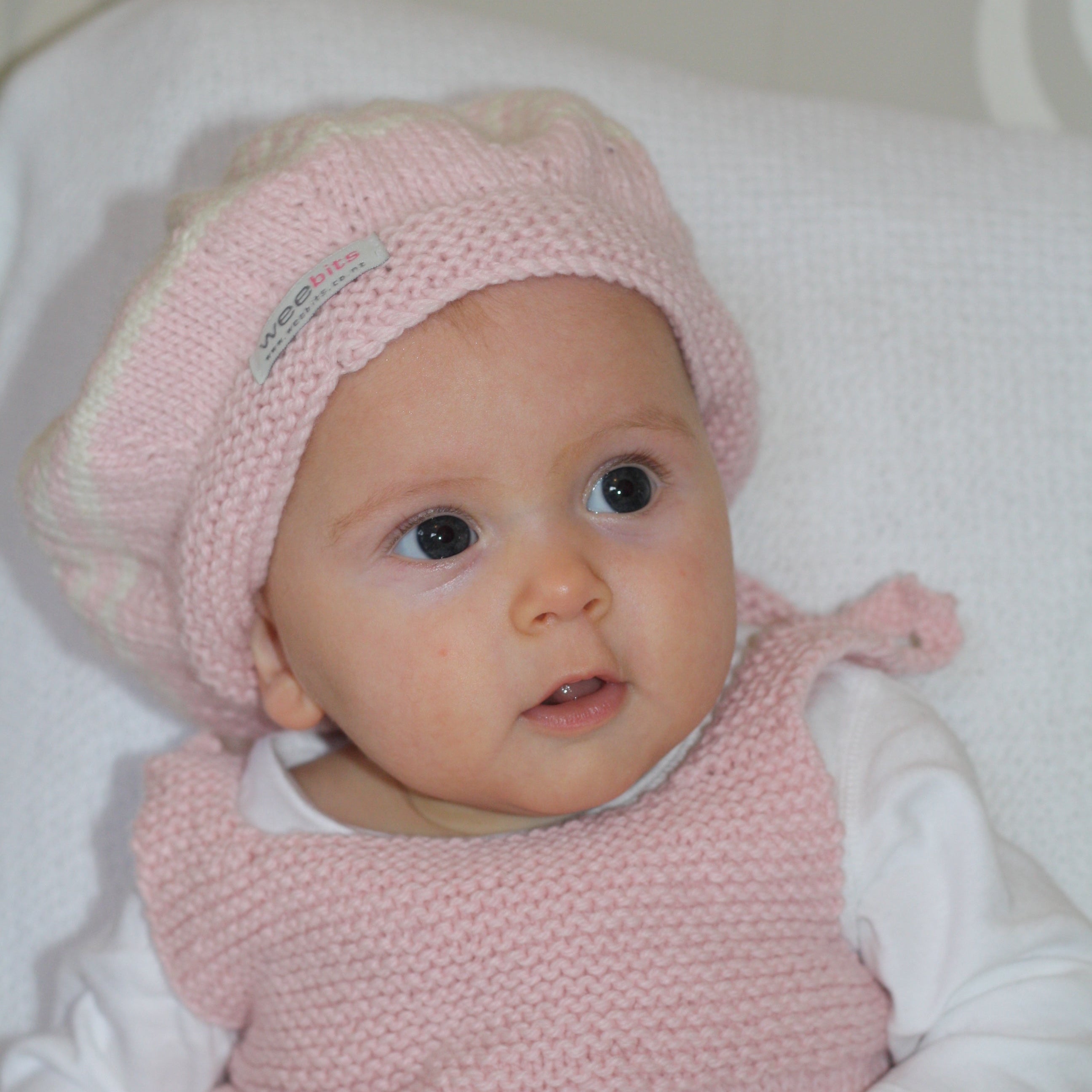 A baby wearing a light pink knit beret and matching pink knit dress over a white long-sleeve shirt, lying on a white textured blanket.