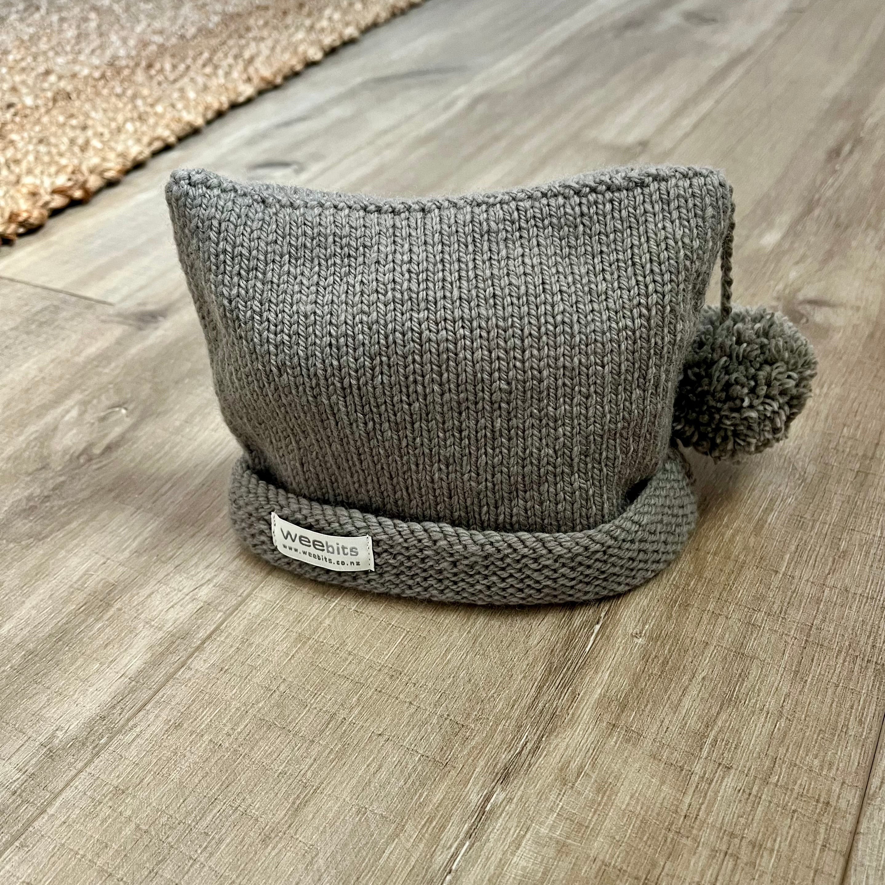 A gray knitted beanie hat with a pom-pom and a small white brand label reading 'Weebits', placed on a light wood floor.