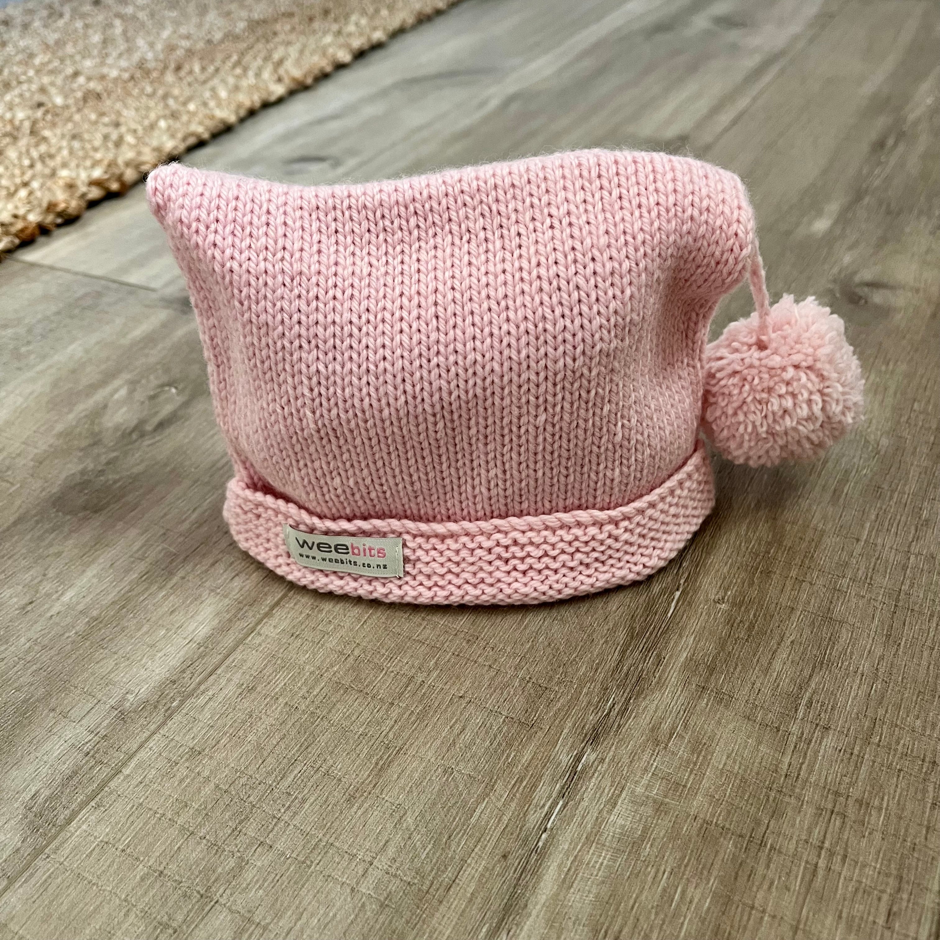 A light pink knit baby hat with a pom-pom and a folded brim, featuring a small white brand label that reads 'weebits', placed on a wood floor.