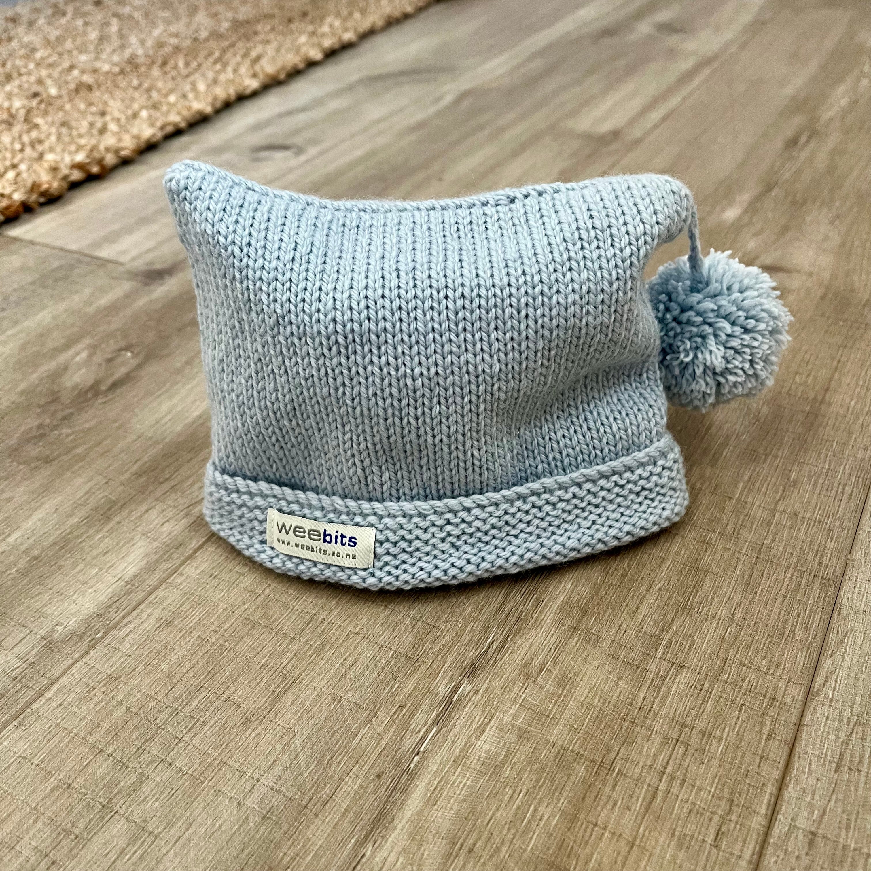 A light blue knit beanie hat with a matching pom-pom and a small white brand label reading 'webbits', placed on a wood floor.