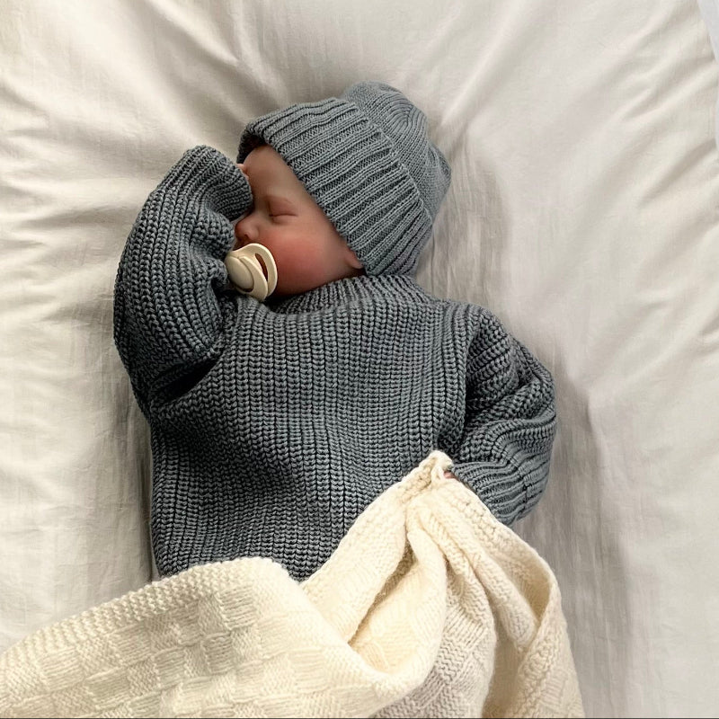 Baby wrapped in a dusky blue jumper and matching beanie with a white blanket, lying on a white surface.
