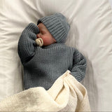 Baby wrapped in a dusky blue jumper and matching beanie with a white blanket, lying on a white surface.