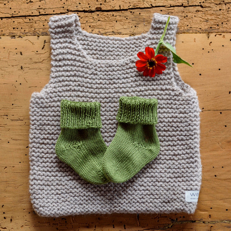 Knitted baby outfit with a oatmeal tank, moss socks, and a red flower on a wooden surface.
