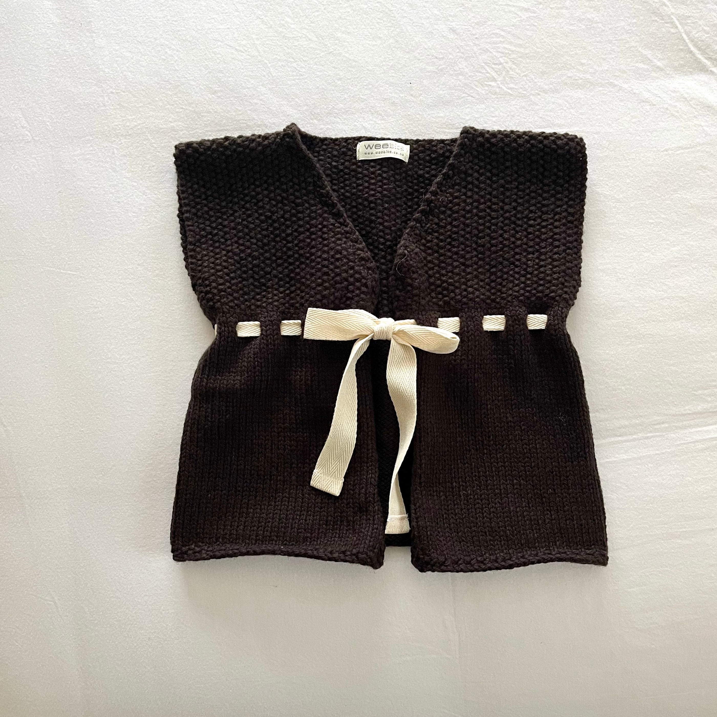 A dark brown knit vest with a cream ribbon belt and bow, laid flat on a white fabric background.
