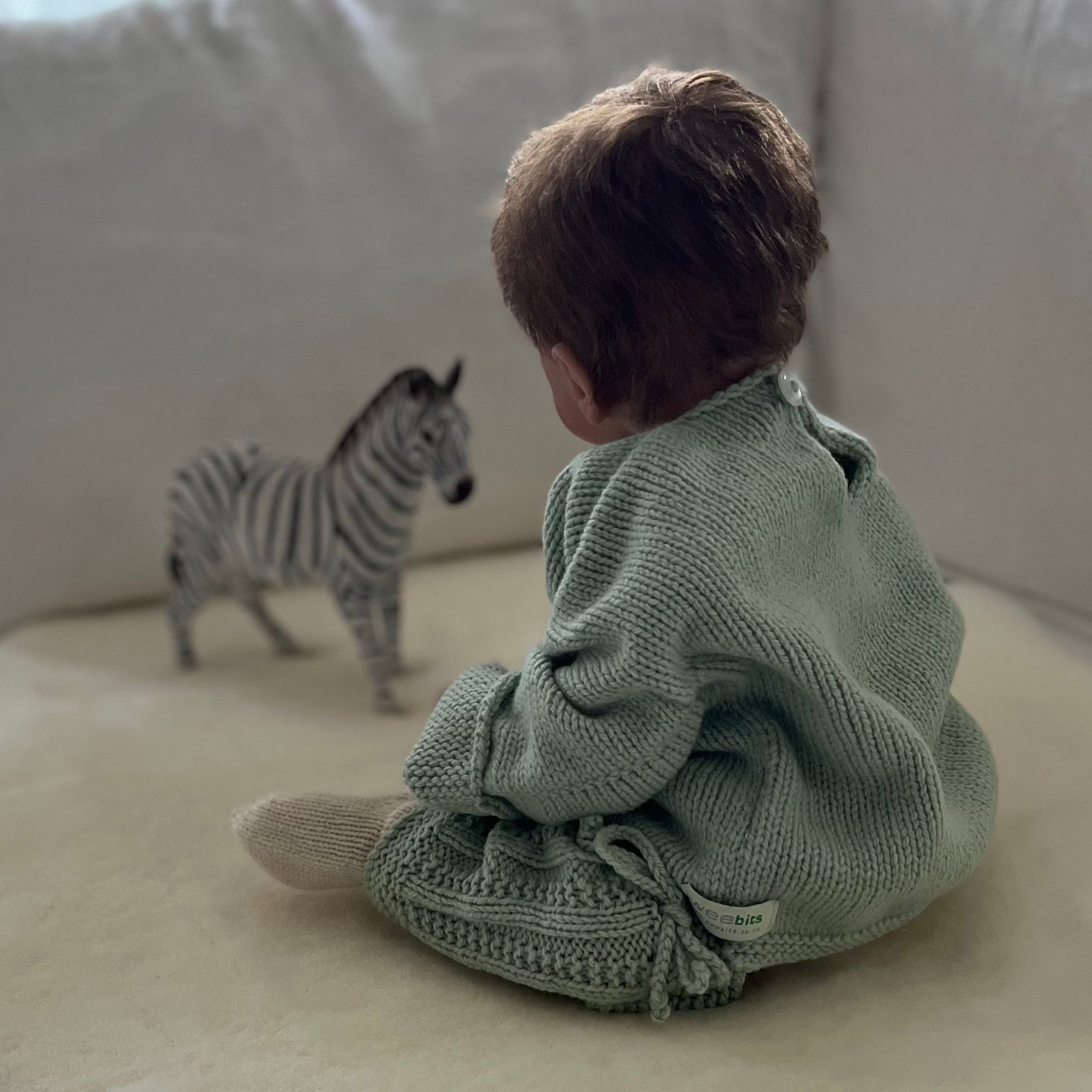 A baby sits on a cream sheepskin rug in a playpen, wearing a sage green knitted jersey and legwarmers. A small zebra toy is placed in front of the baby