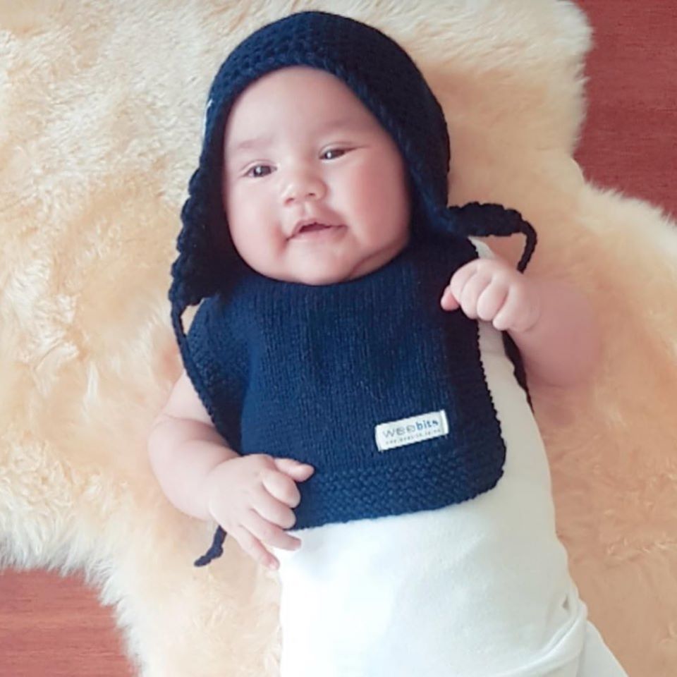 A baby wearing a navy blue knit bonnet and bib set, lying on a cream-colored  rug. The bib features a small white label with the brand name 'weebits'.