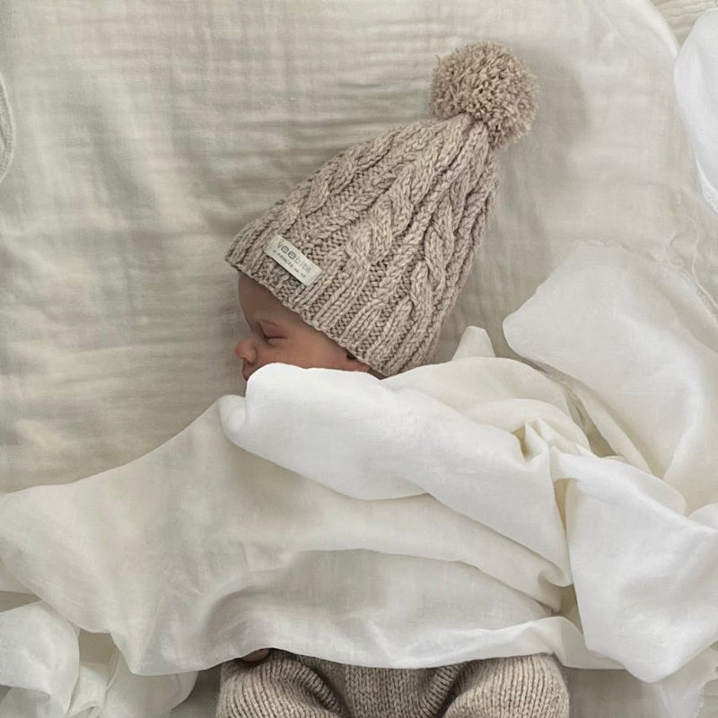 Newborn baby wrapped in a white blanket wearing a oatmeal cable knit hat with a pom-pom.