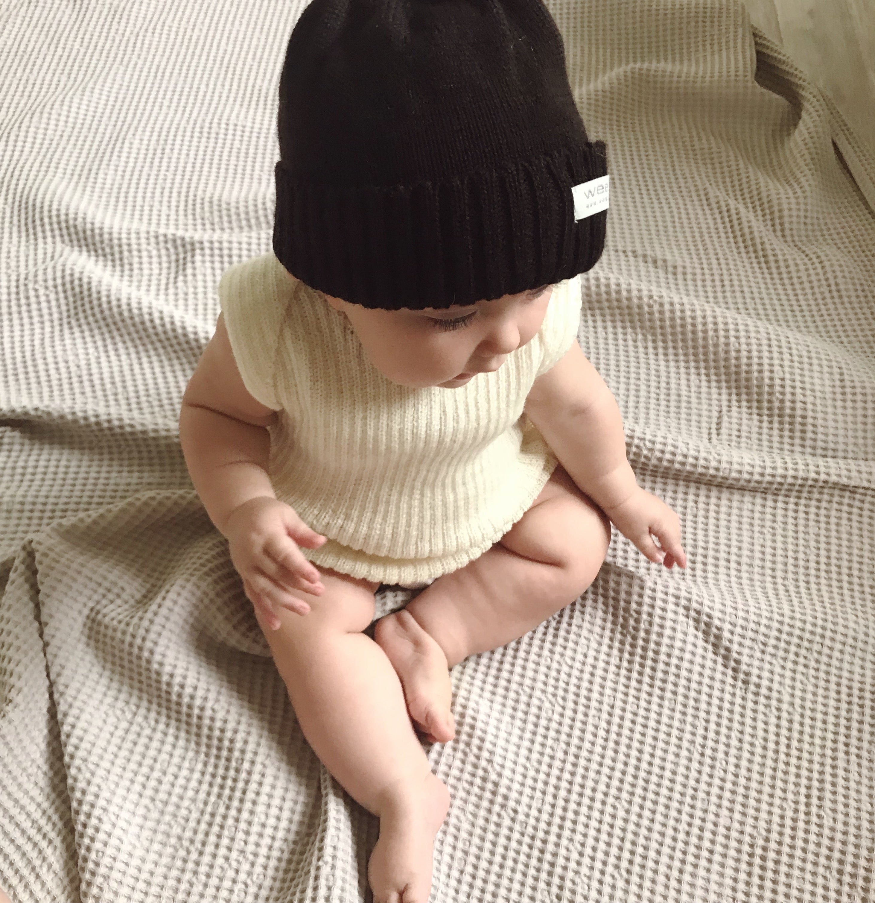 Baby wearing a brown knit hat and cream-colored vest sitting on a textured beige blanket.