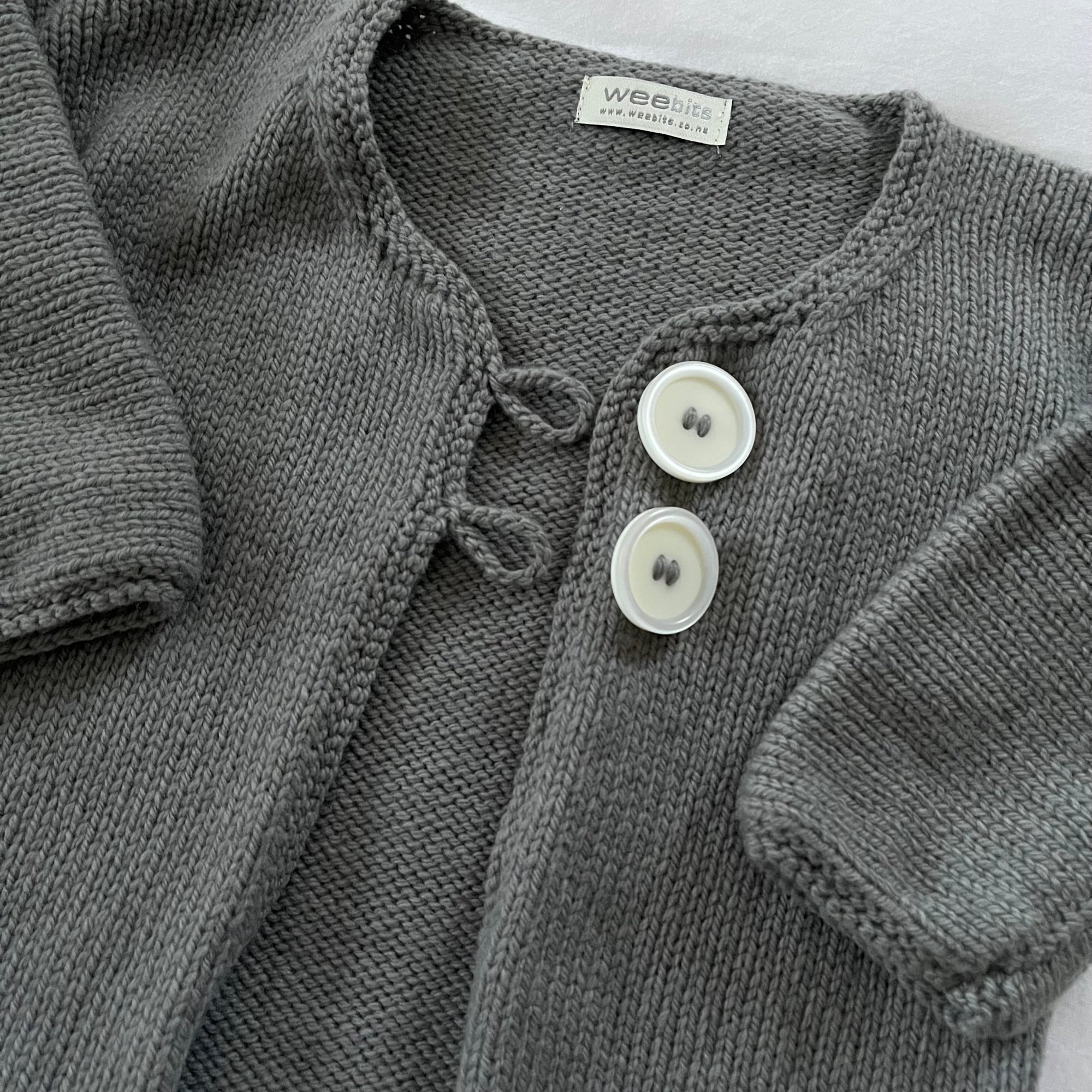 Close-up of a gray knit cardigan with two white buttons, showing the ribbed texture and a visible Weebits brand label.