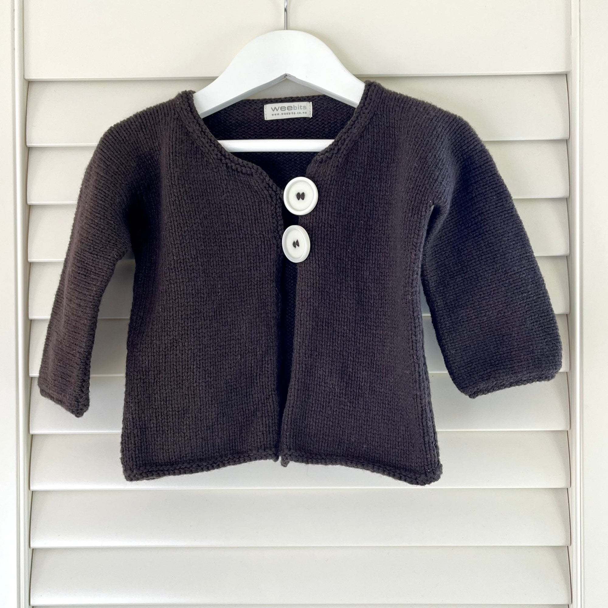 A brown knit baby cardigan sweater with two white buttons, hanging on a white hanger against a white wooden shutter background.