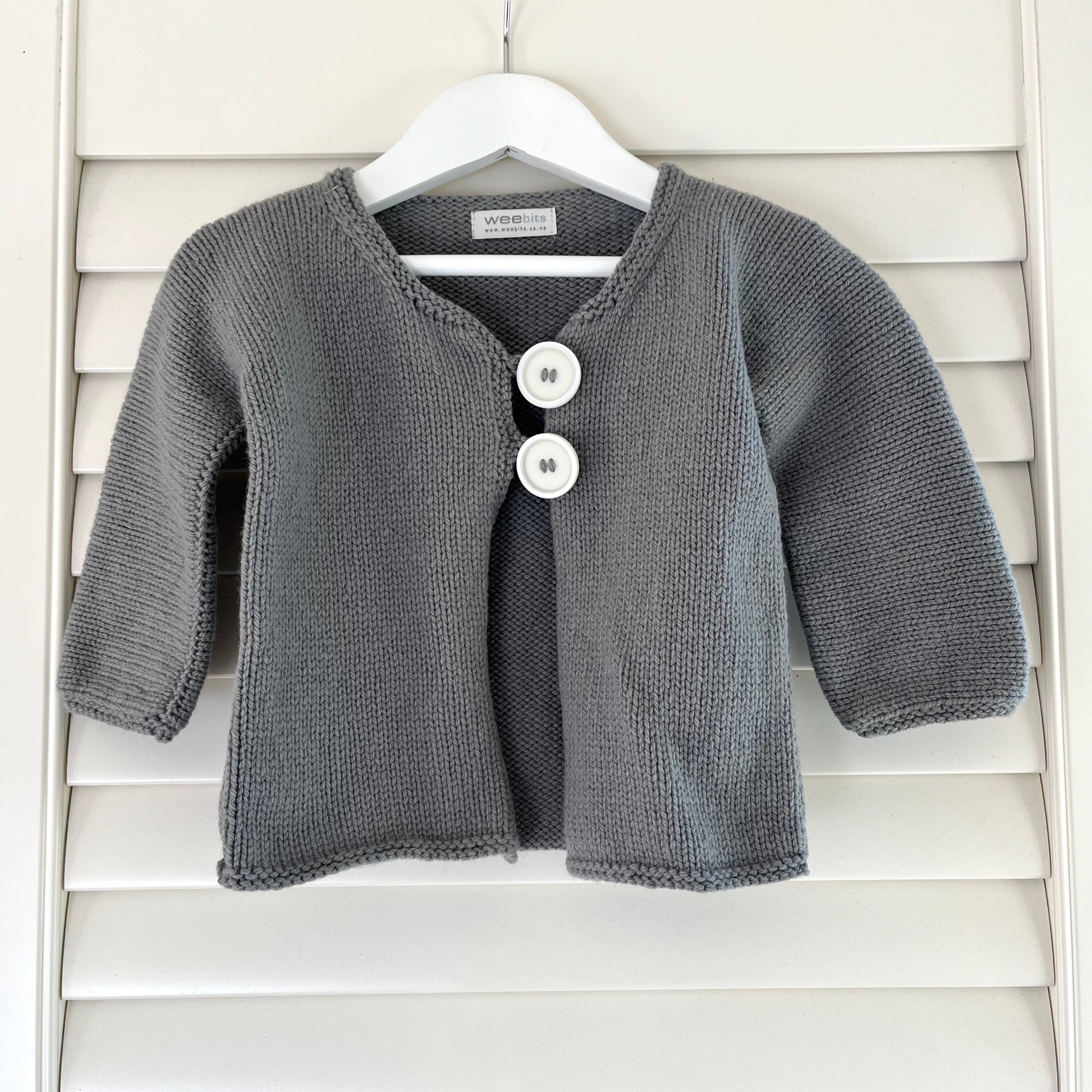 A gray knit baby cardigan sweater with two white buttons, hanging on a white hanger against a white wooden shutter background.