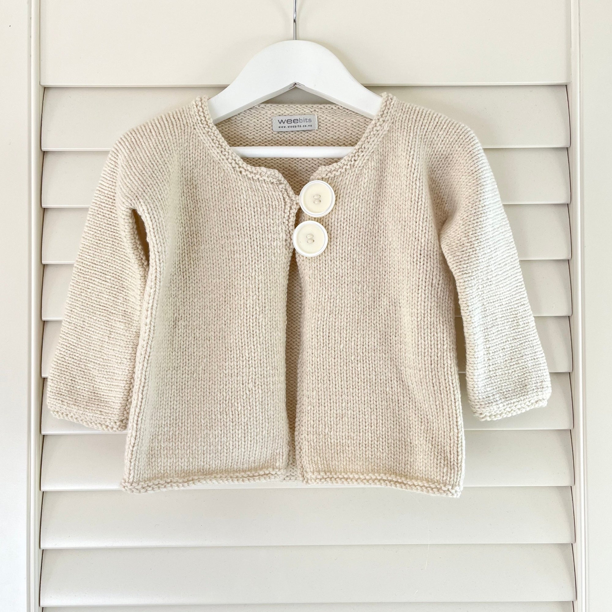A natural knit baby cardigan sweater with two white buttons, hanging on a white hanger against a white wooden shutter background.