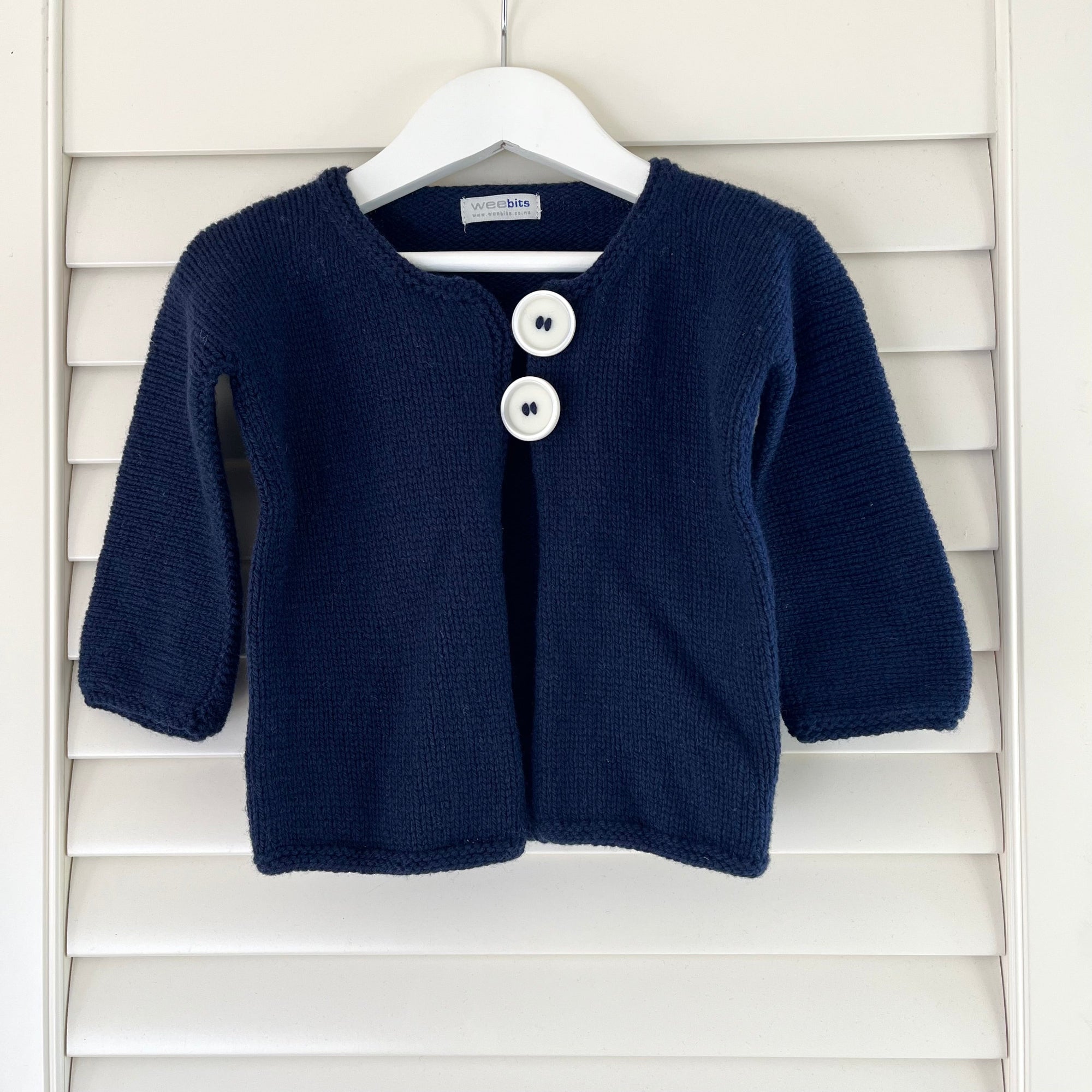 A navy knit baby cardigan sweater with two white buttons, hanging on a white hanger against a white wooden shutter background.