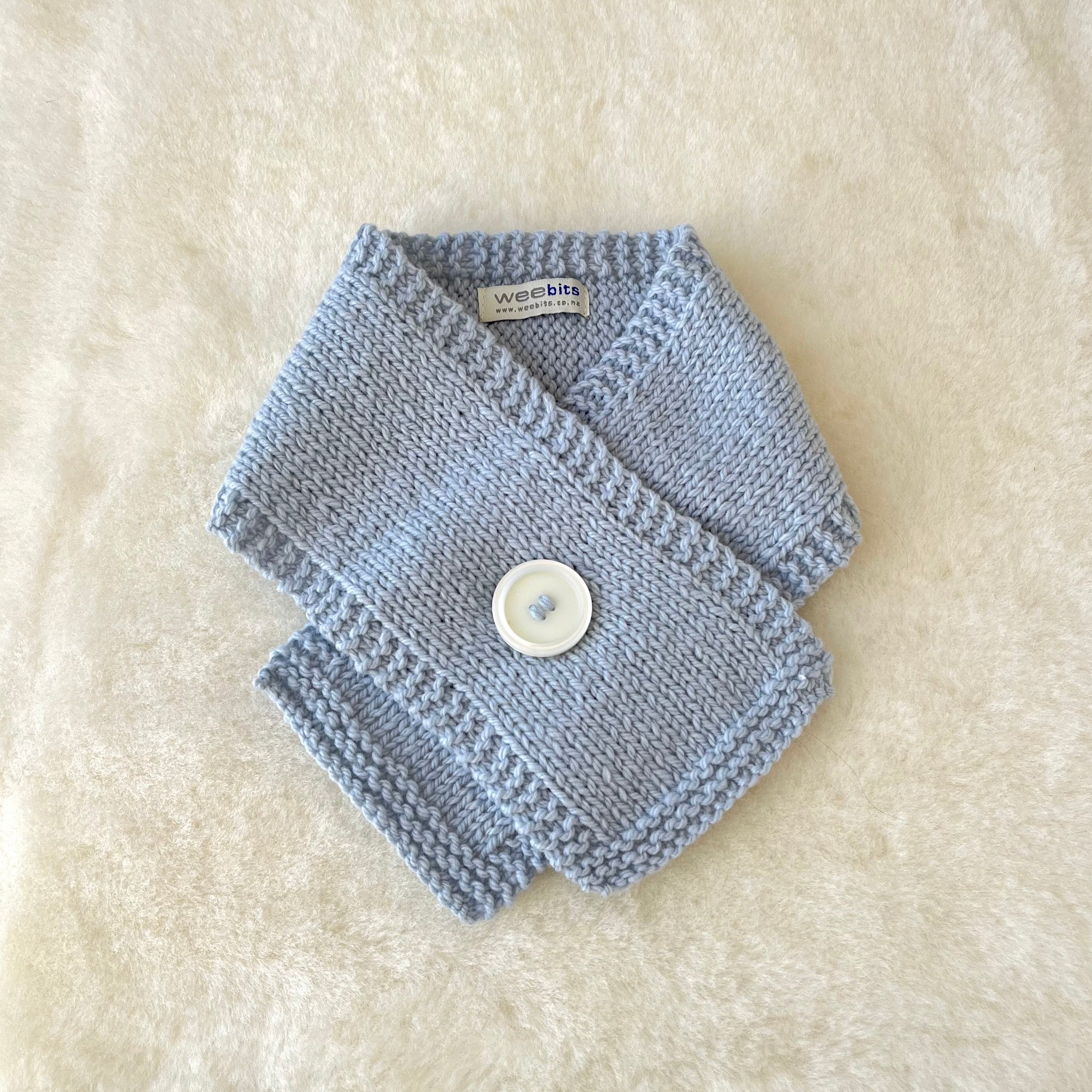 A blue knitted baby scarf with a white button closure, laid flat on a white textured faux fur background.