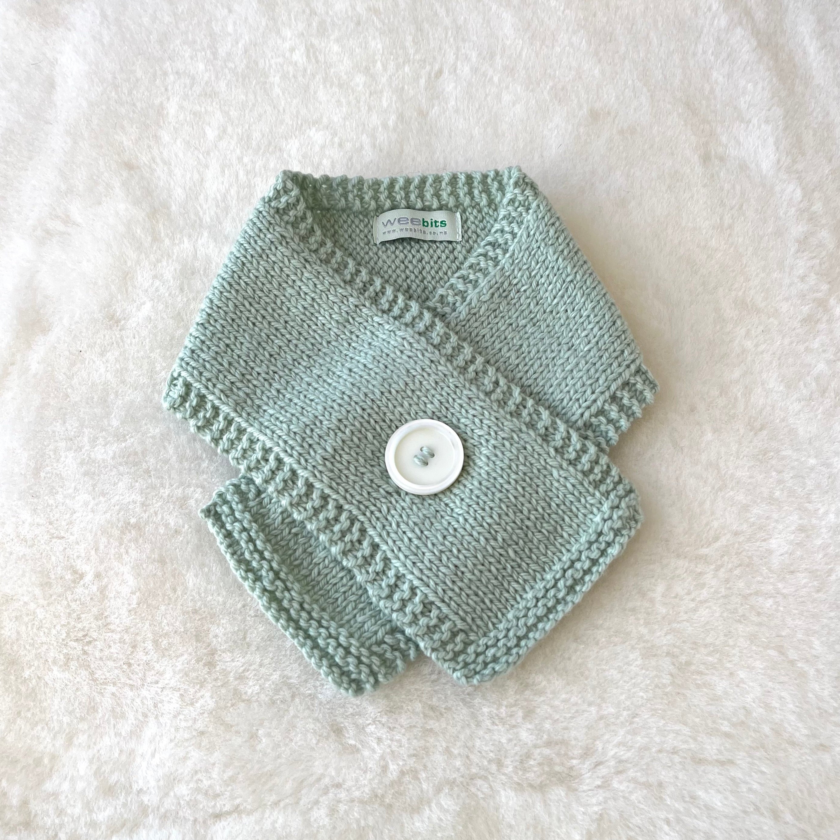 A light sage green knitted baby scarf with a white button closure, laid flat on a white textured faux fur background.