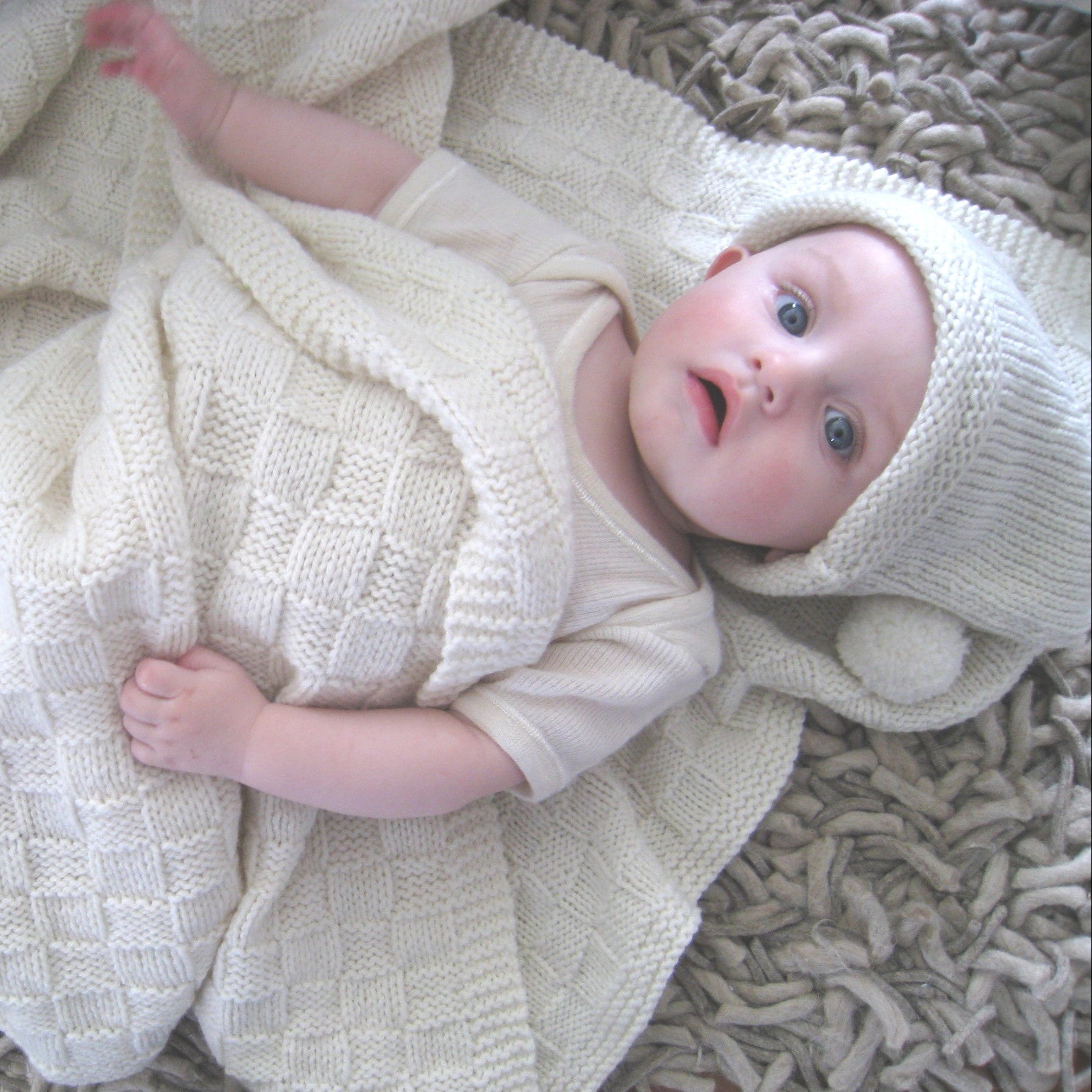 A baby wrapped in a cream-colored knit blanket and matching hat, lying on a textured gray shag rug.