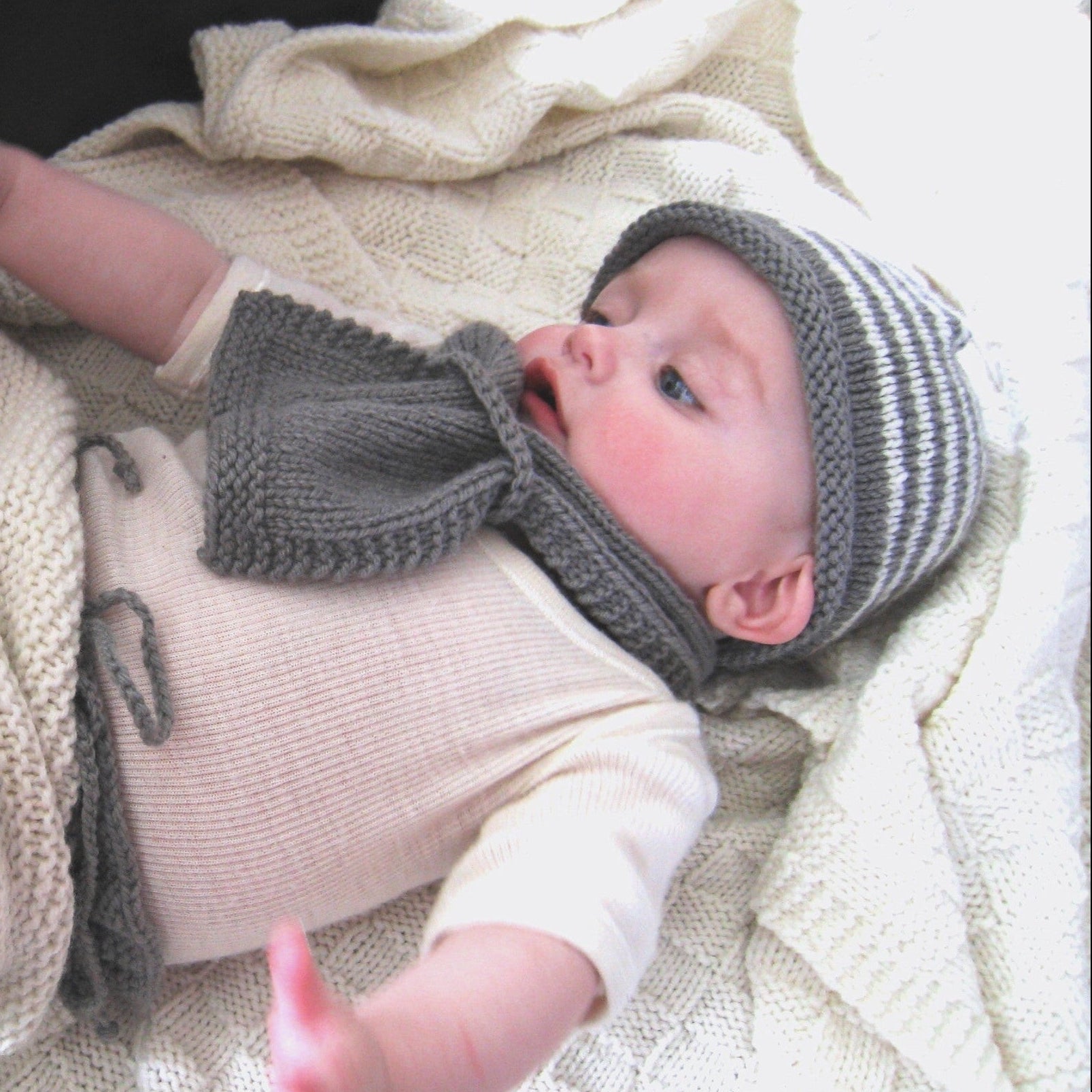 A baby lying on a cream knit blanket, wearing a gray and white striped knit beanie with an attached gray neck warmer and a cream ribbed bodysuit.