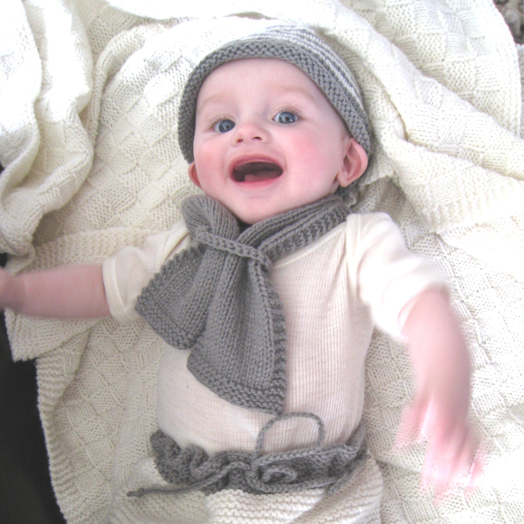 A baby lying on a cream knit blanket, wearing a cream short-sleeve romper with a matching gray knit hat and scarf.