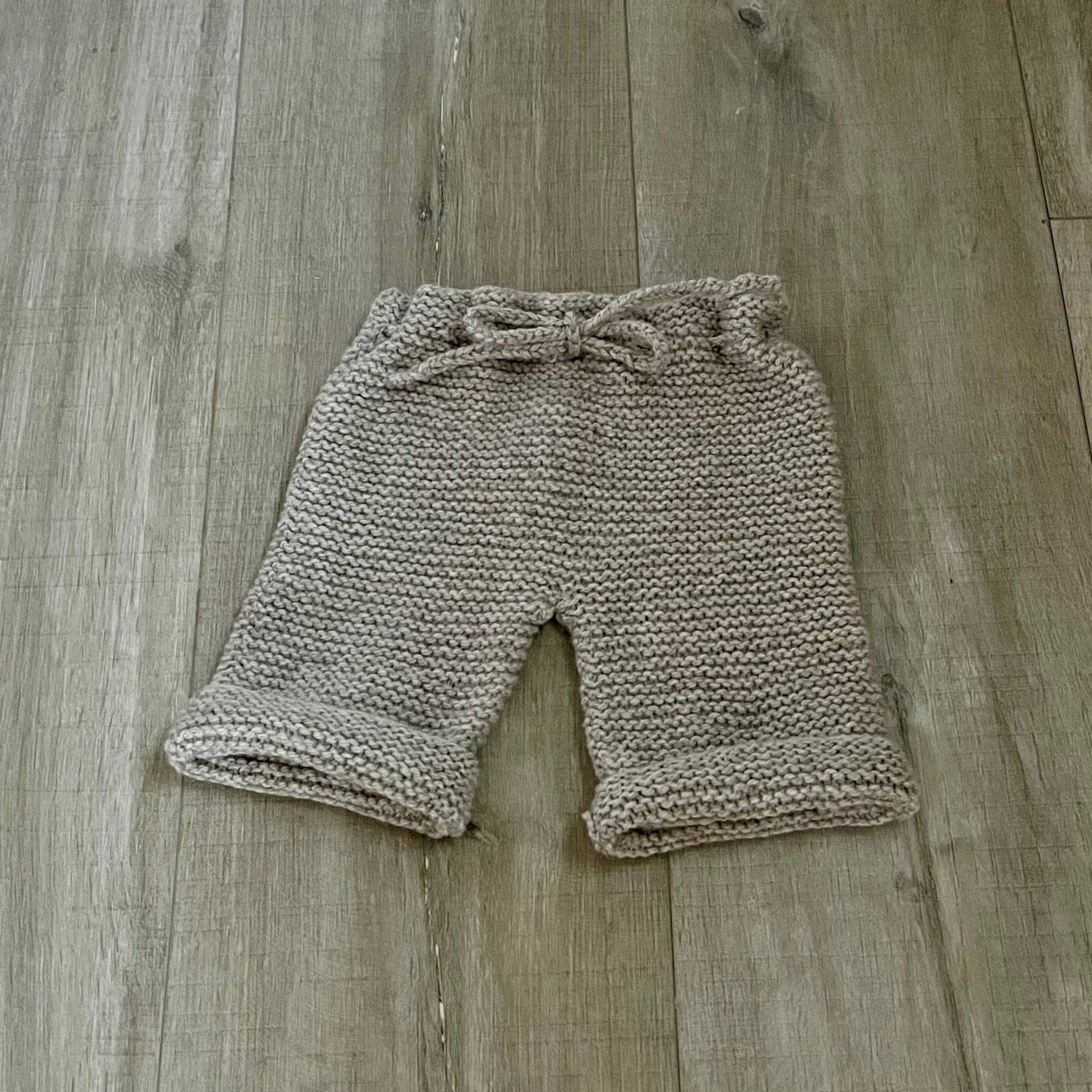 Hand-knit baby shorts in a warm taupe-beige color, featuring a matching drawstring waist and ribbed cuffs, laid flat on a white fabric background.