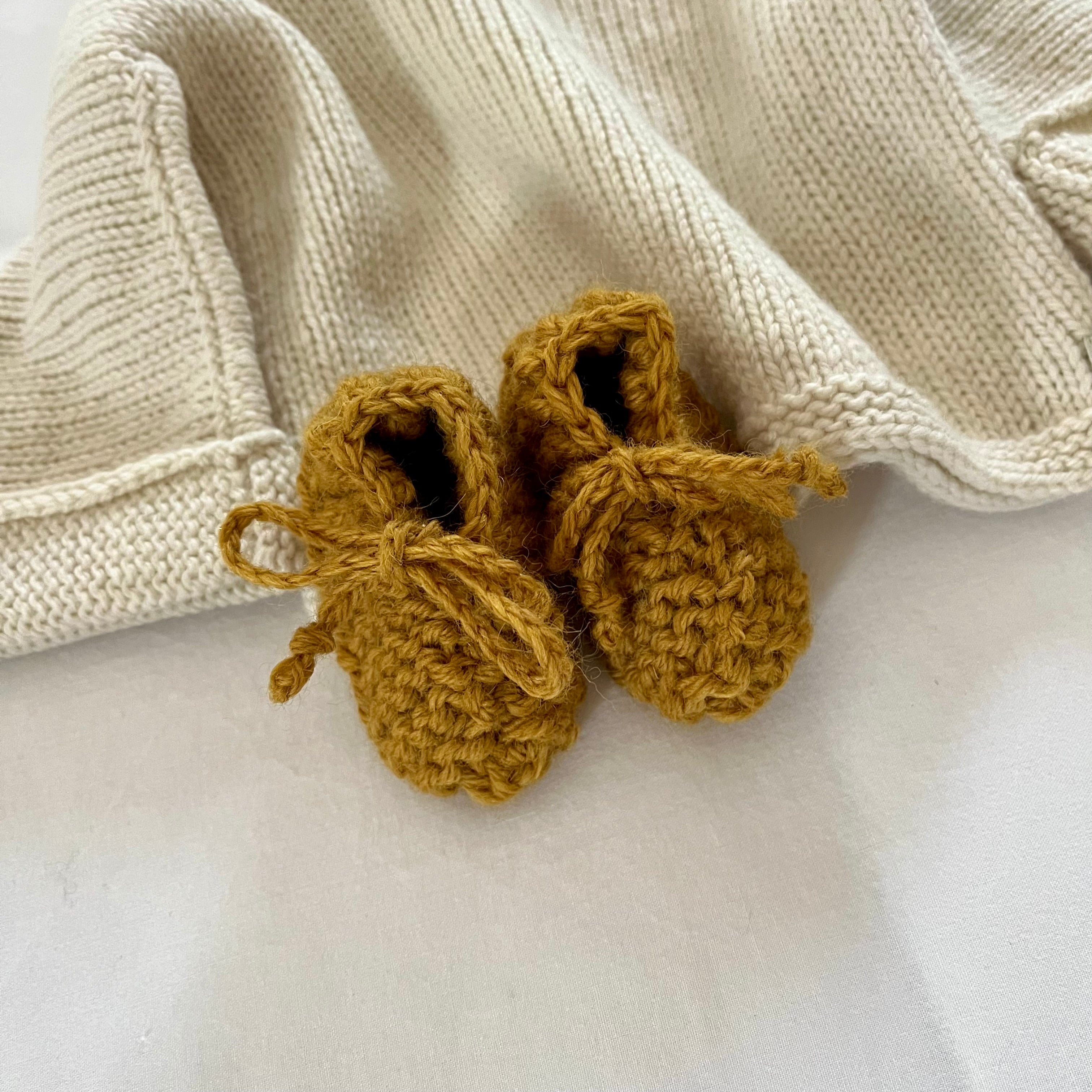 Pair of mustard yellow knitted baby booties on a white surface with a beige textured sweater.