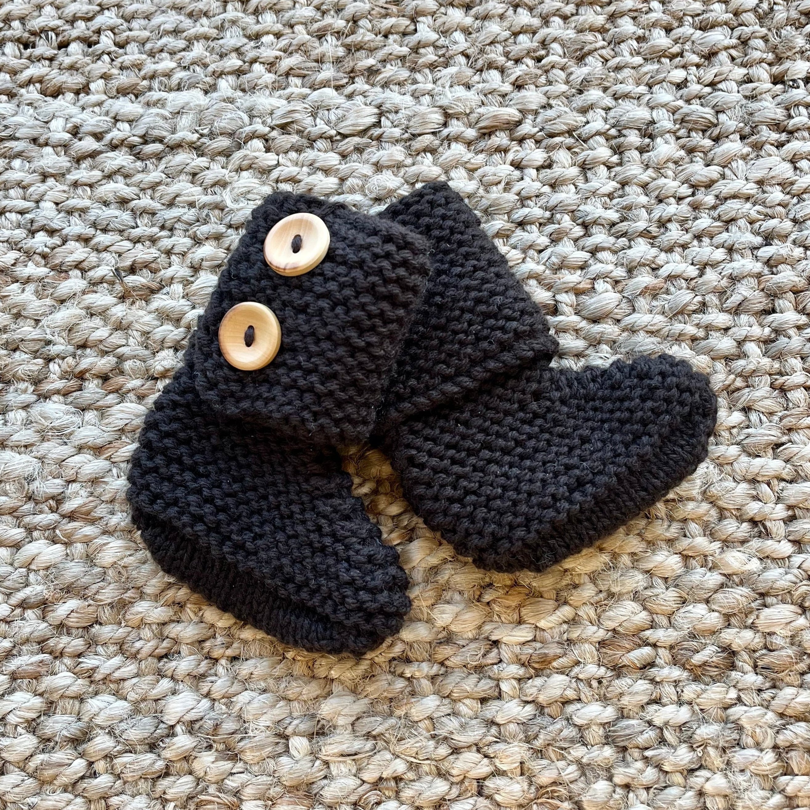 A pair of chocolate knit booties with two light wood buttons, displayed on a textured beige woven rug.